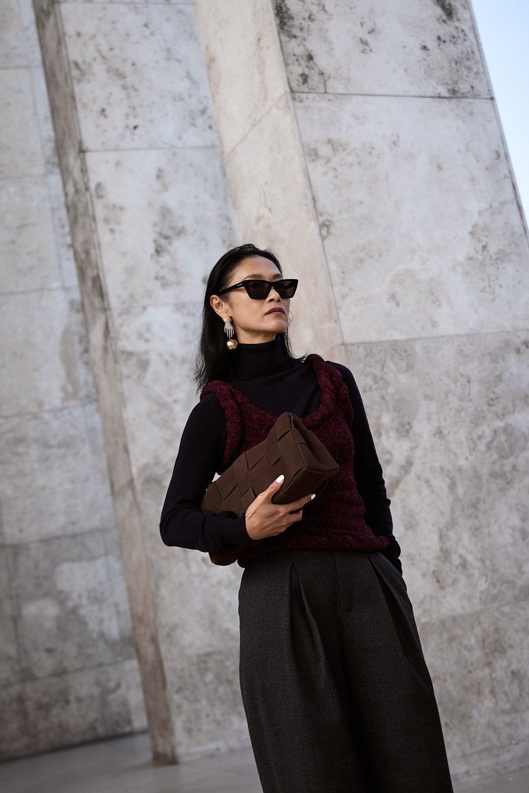 Woman in sunglasses, black polo neck and burgundy knit vest holding a brown clutch, stone wall behind
