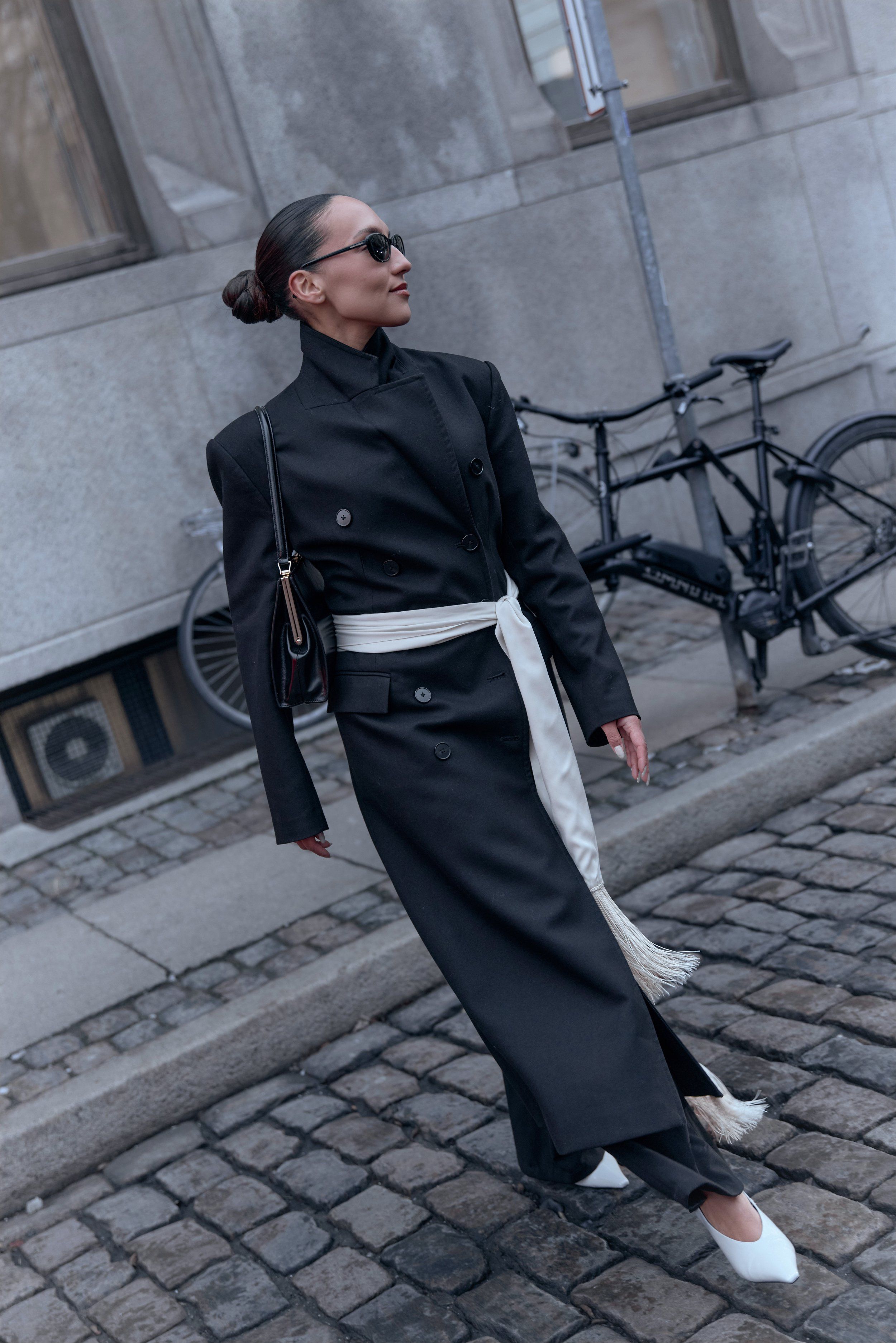 Copenhagen Fashion Week street style: long black coat with white sash and white pointed shoes on cobbles