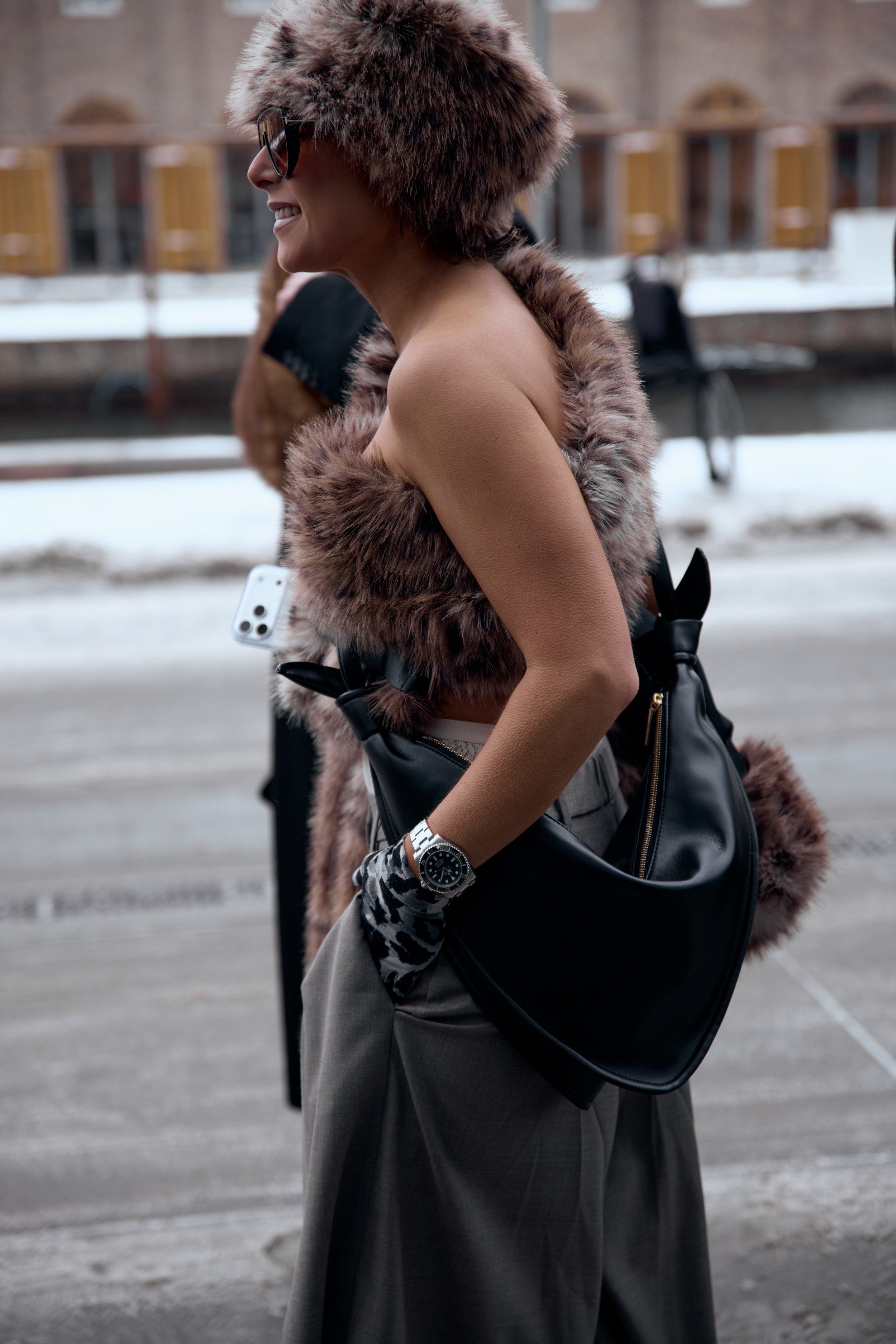 Woman in a brown feather-trim strapless top and a black crescent bag over grey trousers by a snowy canal