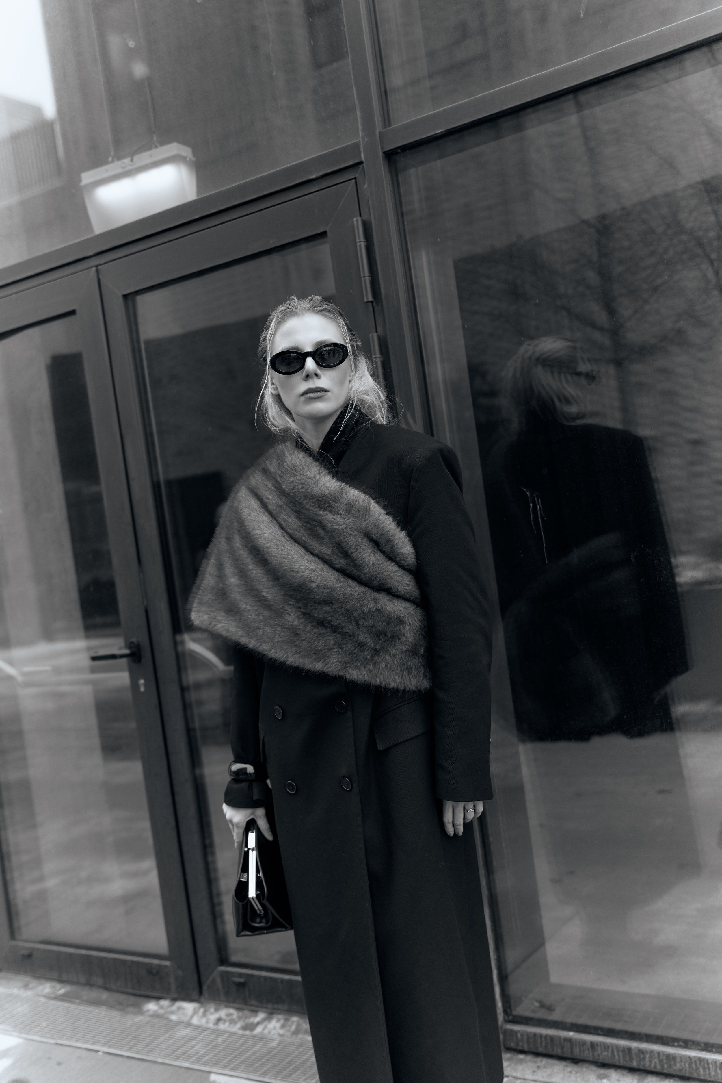 Black-and-white portrait of a woman in sunglasses, dark long coat and grey knit shawl by glass doors