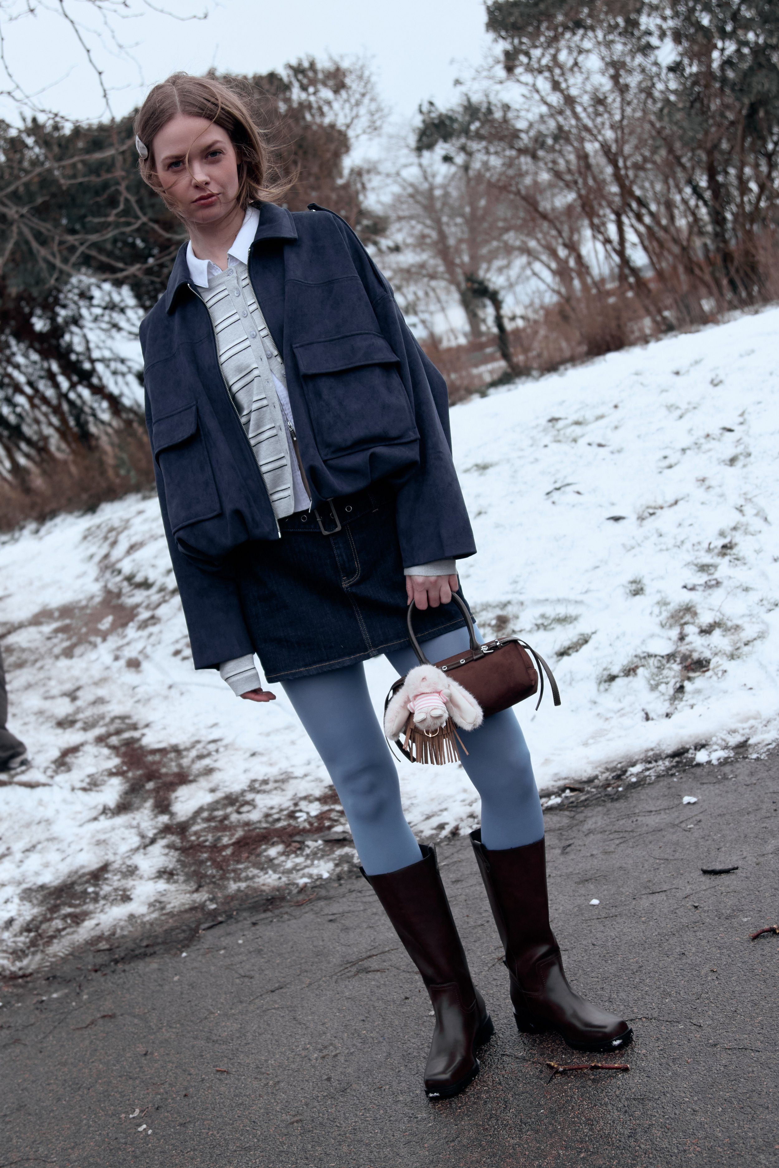 Woman in a navy utility jacket, denim skirt, blue tights and brown boots, snow behind, small bag