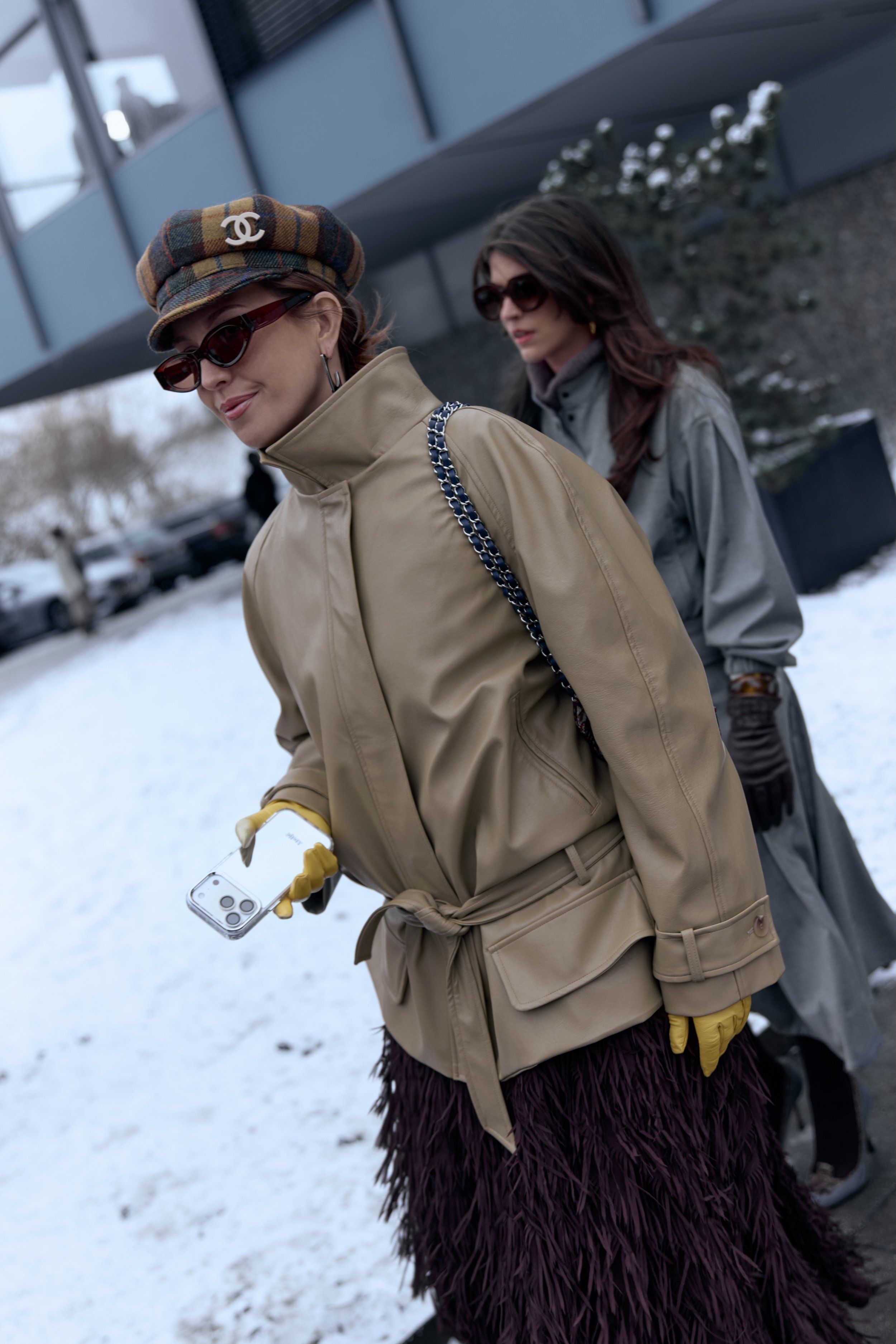Woman in a beige belted jacket and plaid cap, yellow gloves, a feathered burgundy skirt, another guest behind