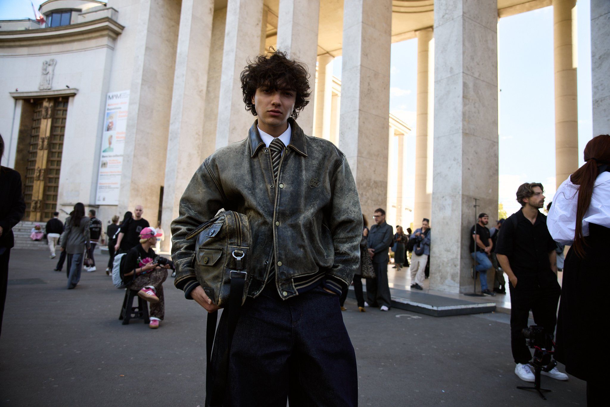 Man in an olive bomber jacket and black trousers in front of columns, crowds in the distance