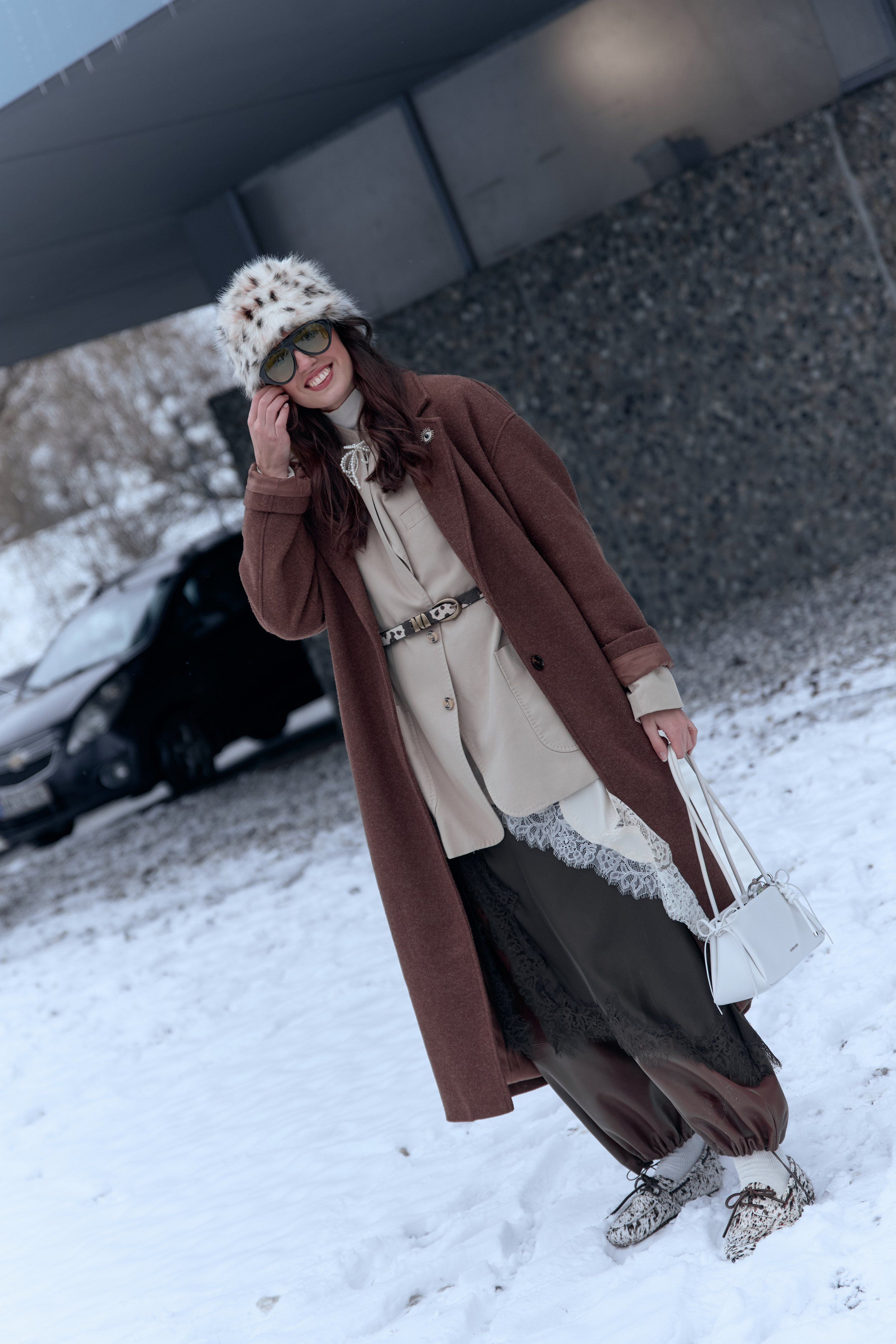 Woman in a patterned bucket hat, brown long coat, belted cream top, white bag and sneakers in snow