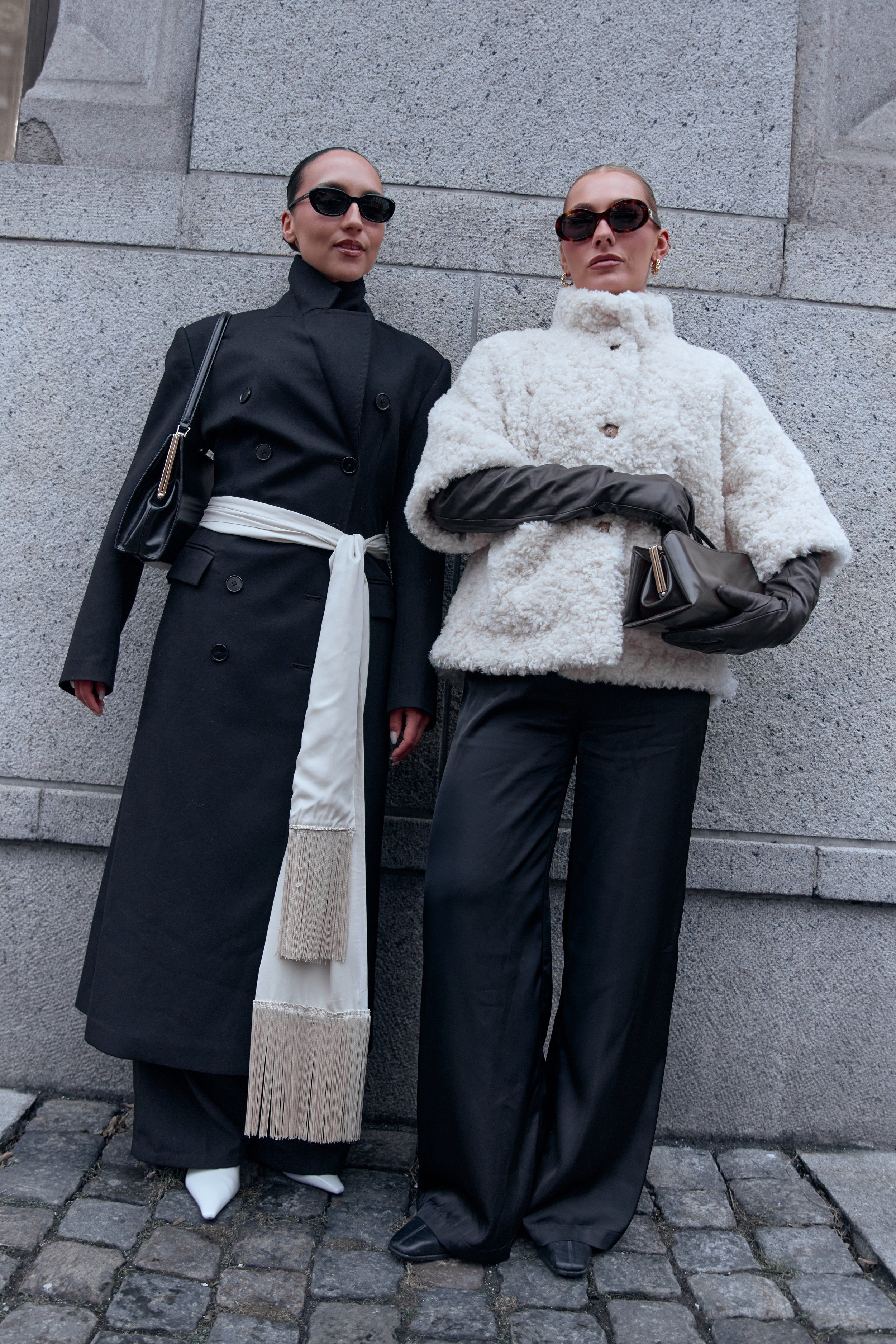 Two guests in sunglasses against a granite wall, one in a black coat with white sash, one in white shearling