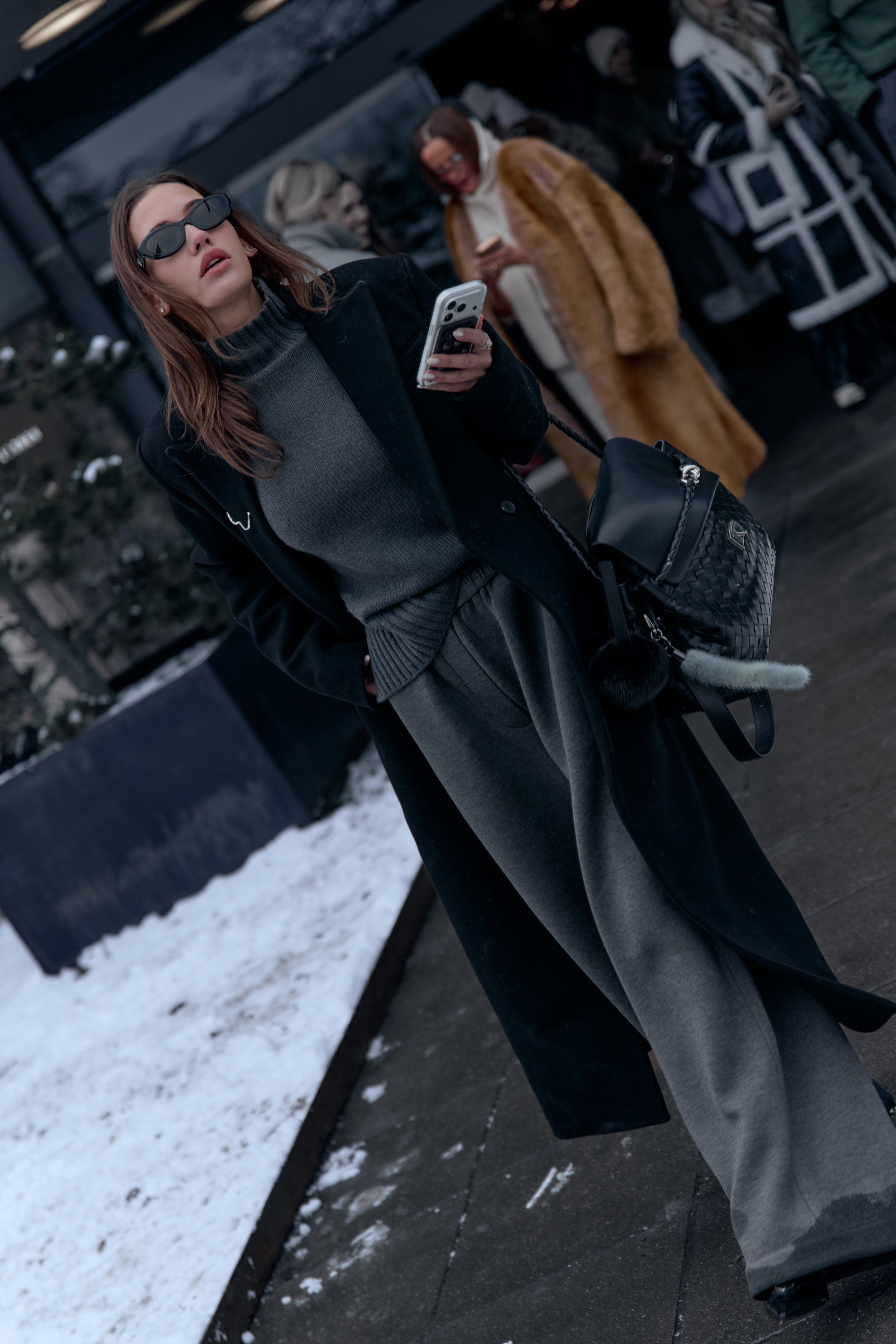 Woman in sunglasses and a black coat over a grey knit and wide trousers, holding a phone, backpack on shoulder
