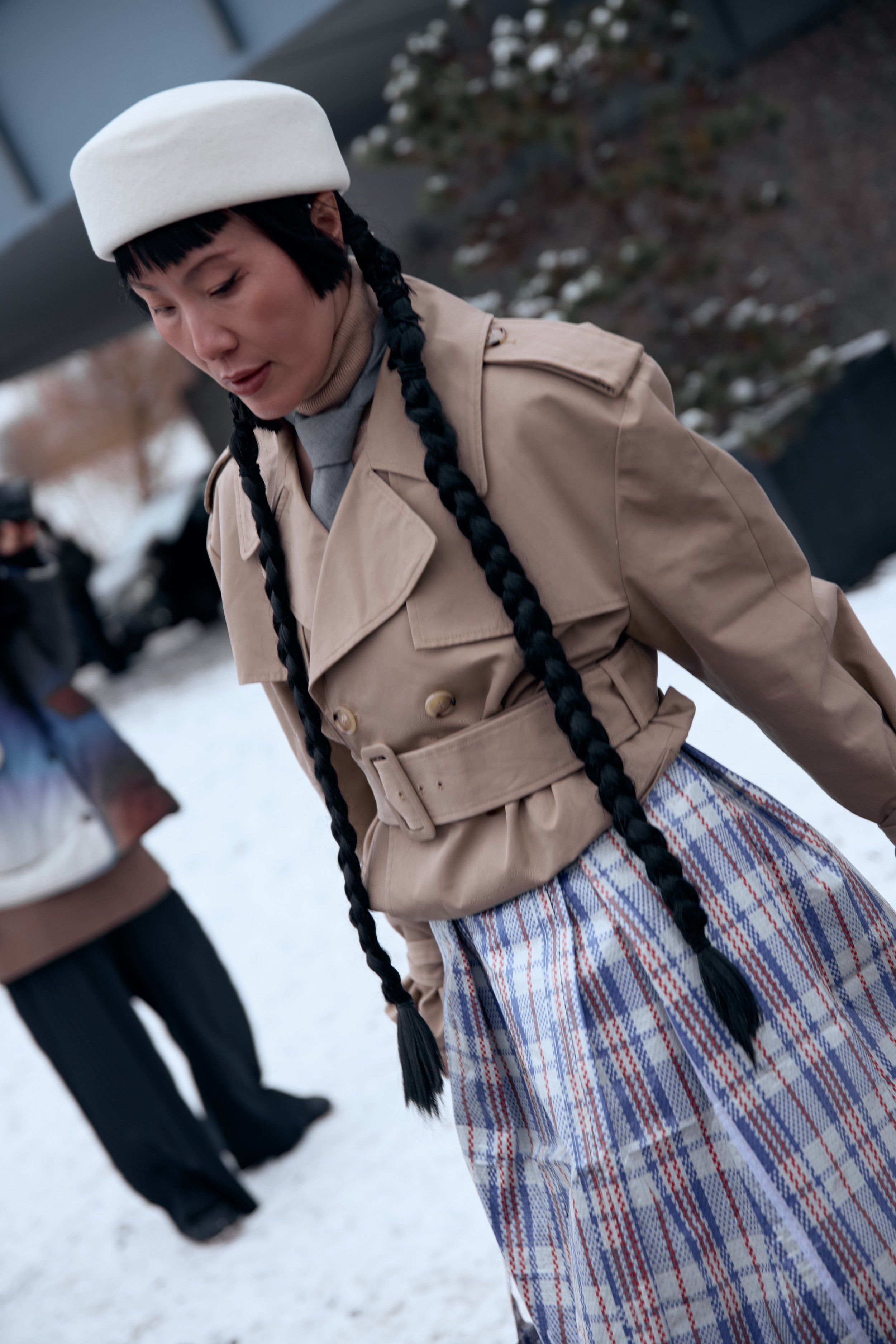 Woman with long black braids, white pillbox hat, beige trench and plaid skirt in the snow