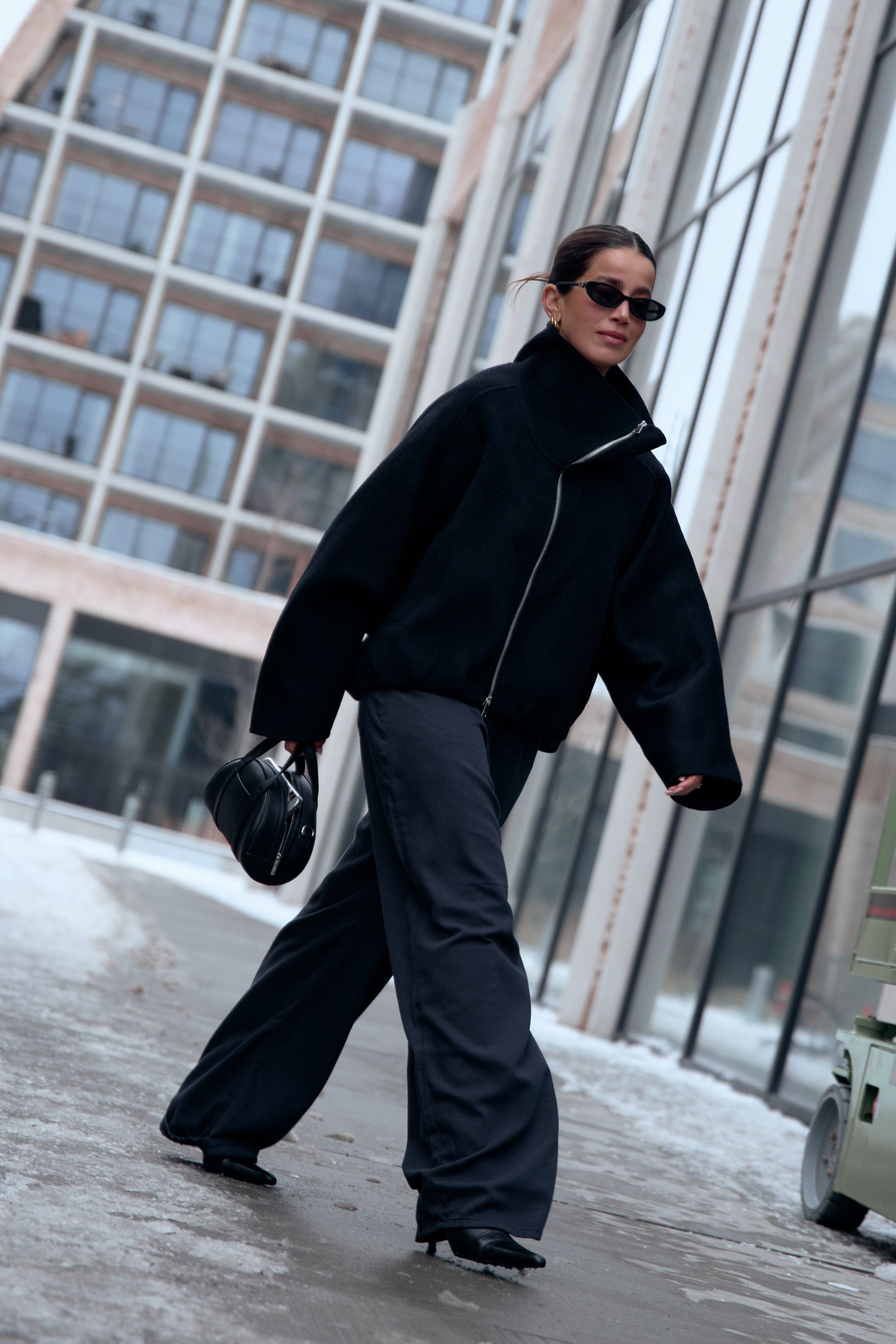 Woman in sunglasses, black jacket and wide trousers carrying a black bag, walking on icy pavement