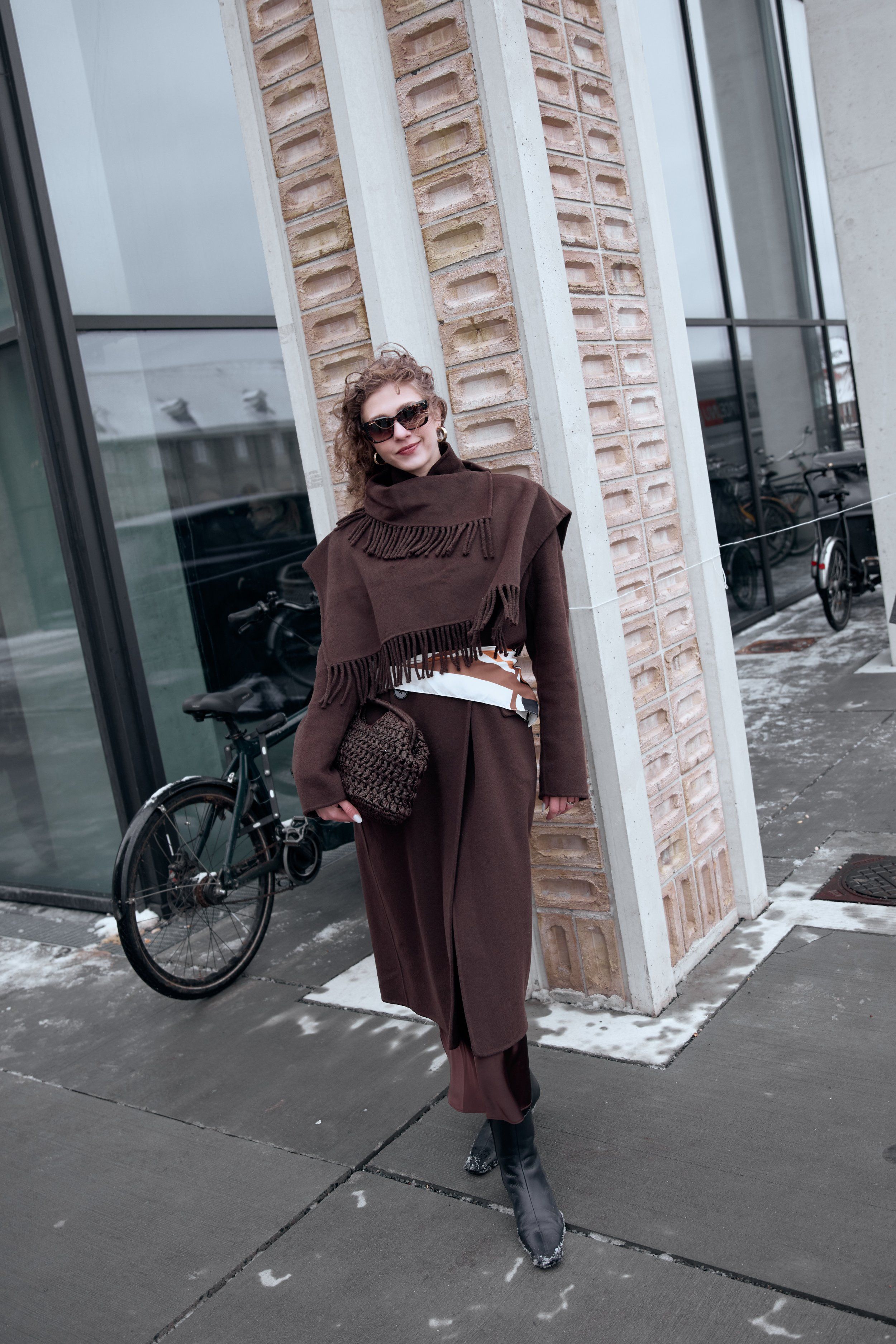 Woman in a brown fringed poncho and long skirt with a woven bag, bicycle and glass building behind