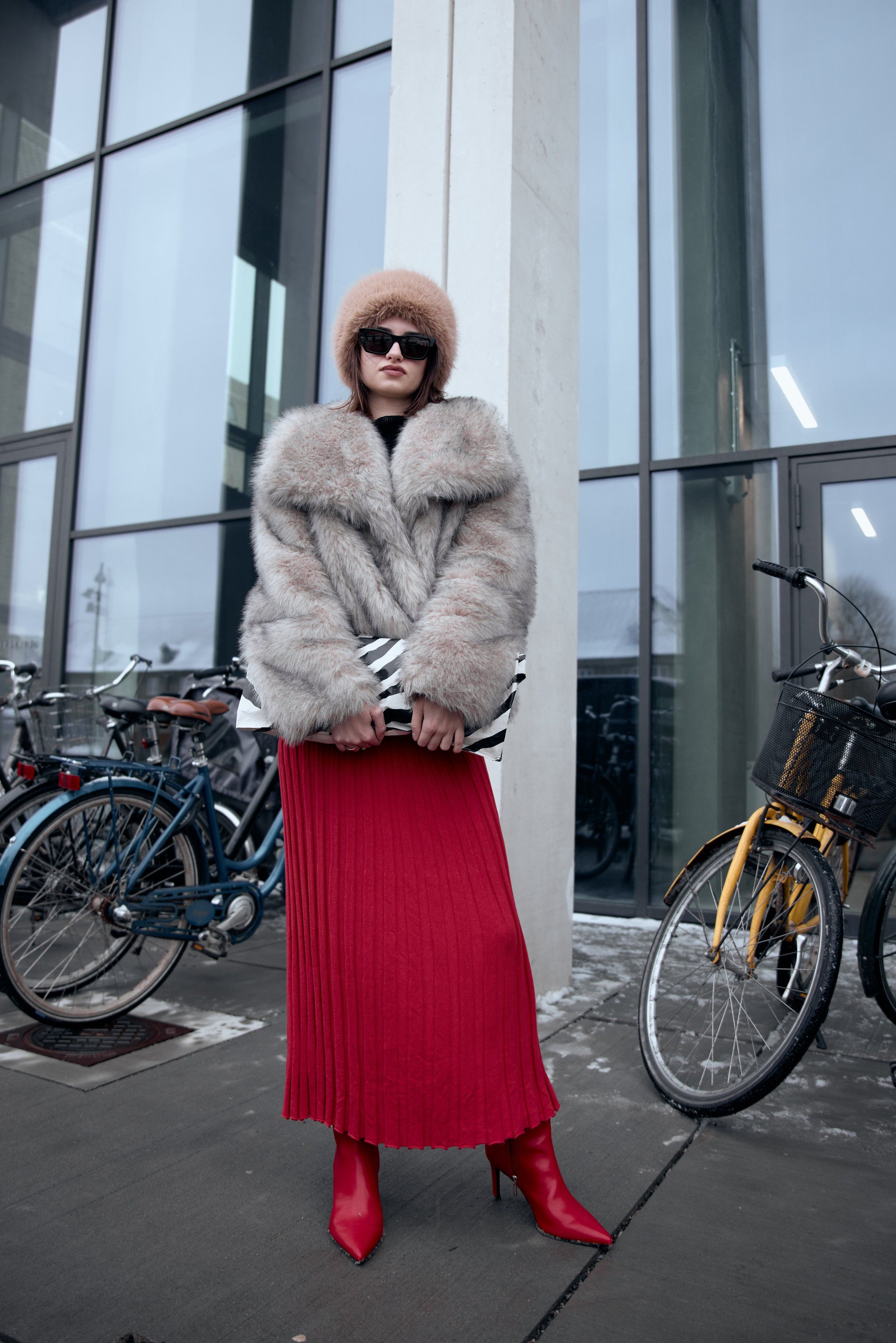 Woman in a grey fur coat, sunglasses and brown hat over a red pleated skirt and red boots, bicycles around