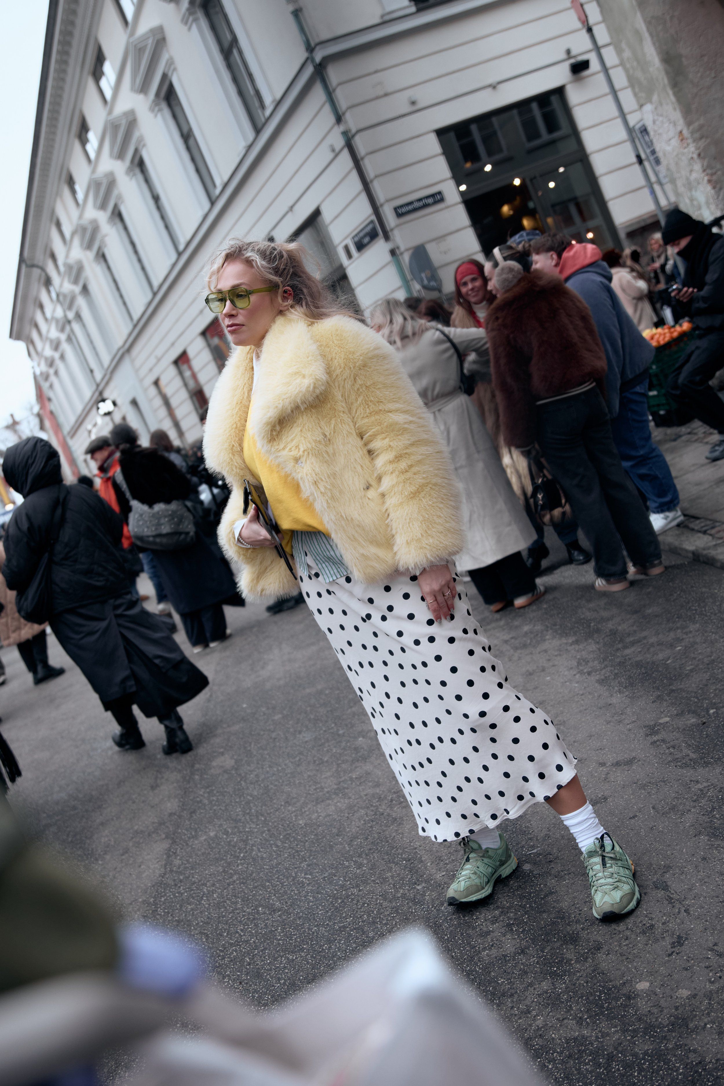 Woman in a yellow fur jacket, polka-dot skirt and green sneakers on a busy street corner