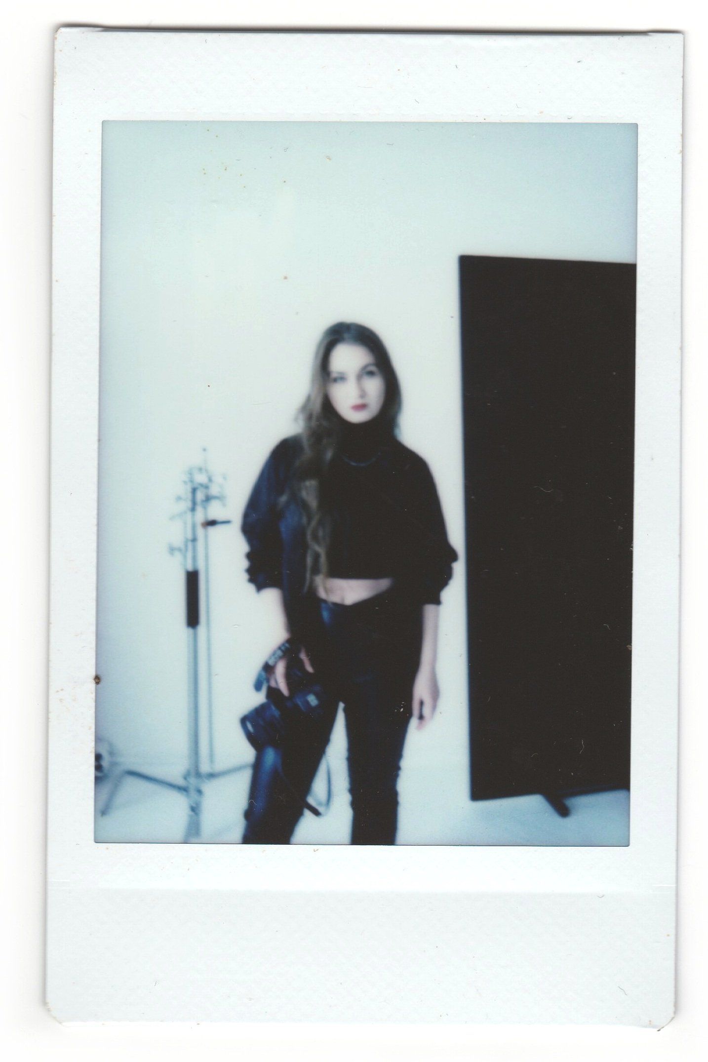 Instant-film portrait of a photographer in black holding a camera beside a lighting stand and black reflector