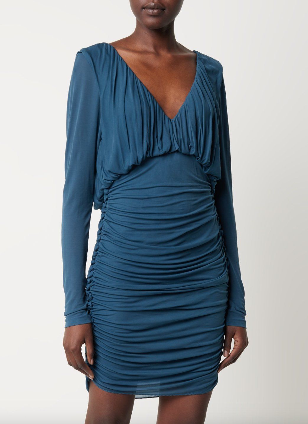 E-commerce shot of a model in a teal ruched long-sleeve mini dress with V-neckline, plain light backdrop