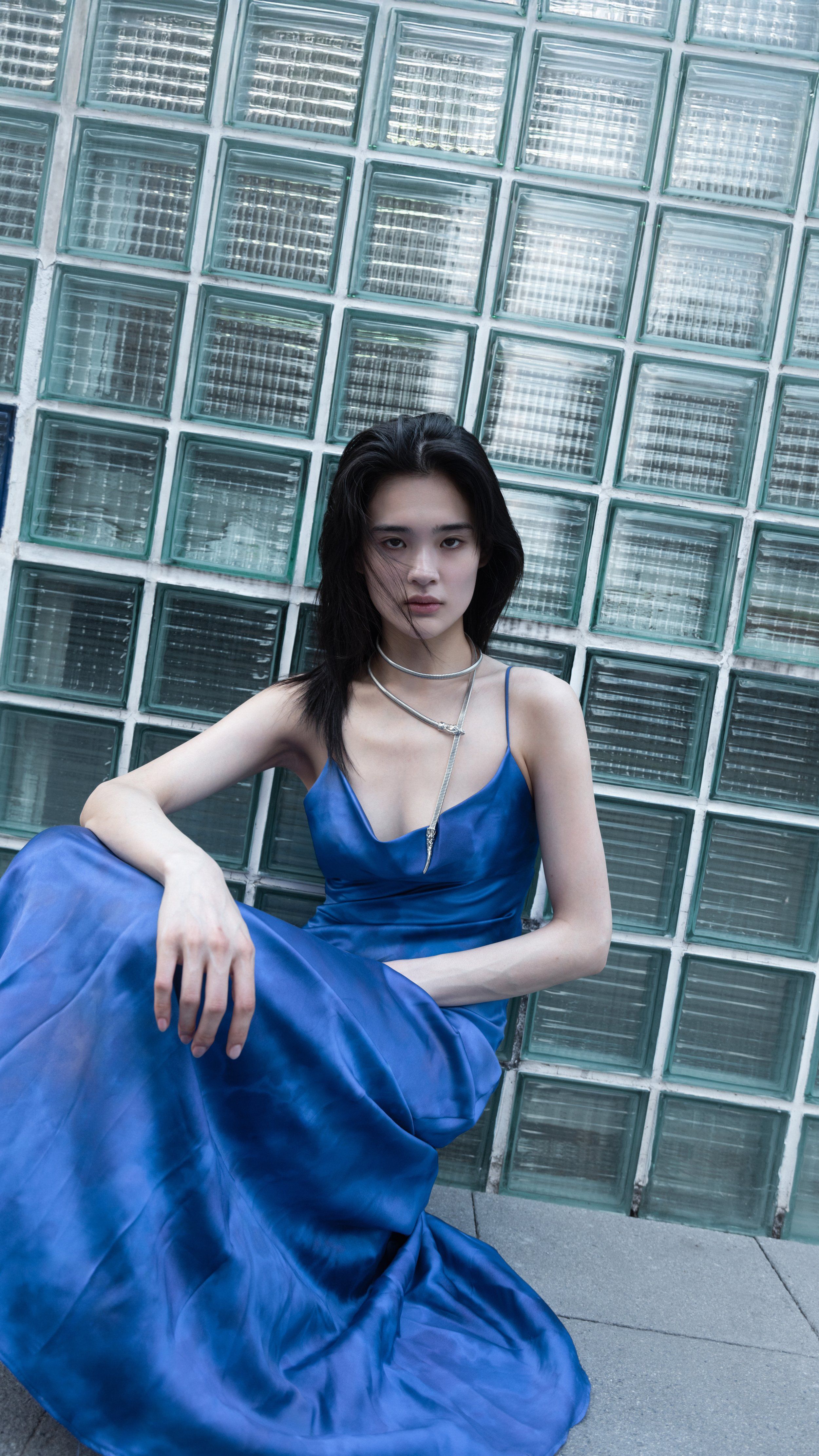 Model seated against a glass-block wall in a cobalt blue satin slip dress, layered necklaces, tall crop