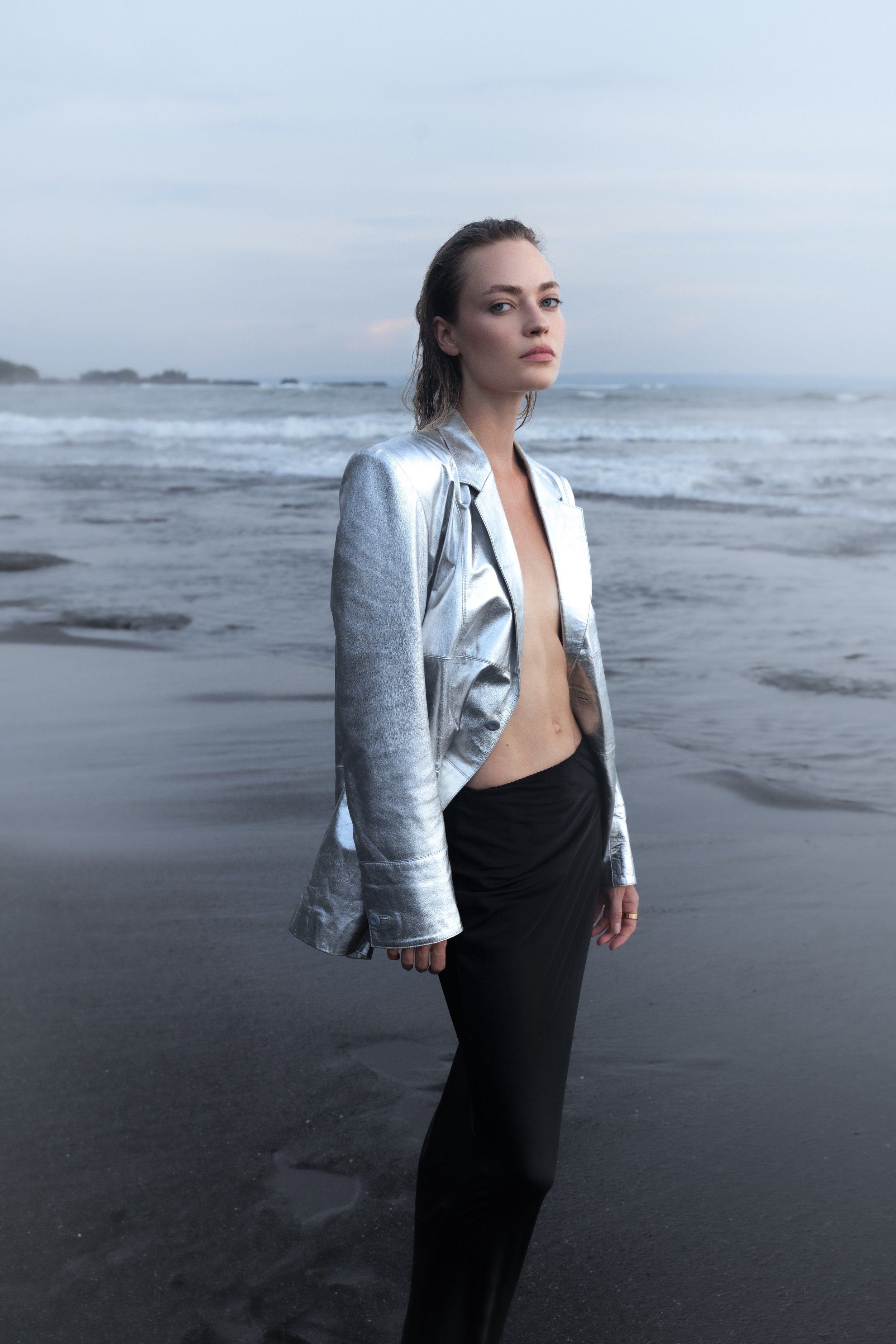 Model on a dark beach at dusk in a silver metallic jacket worn open over black trousers, calm sea behind
