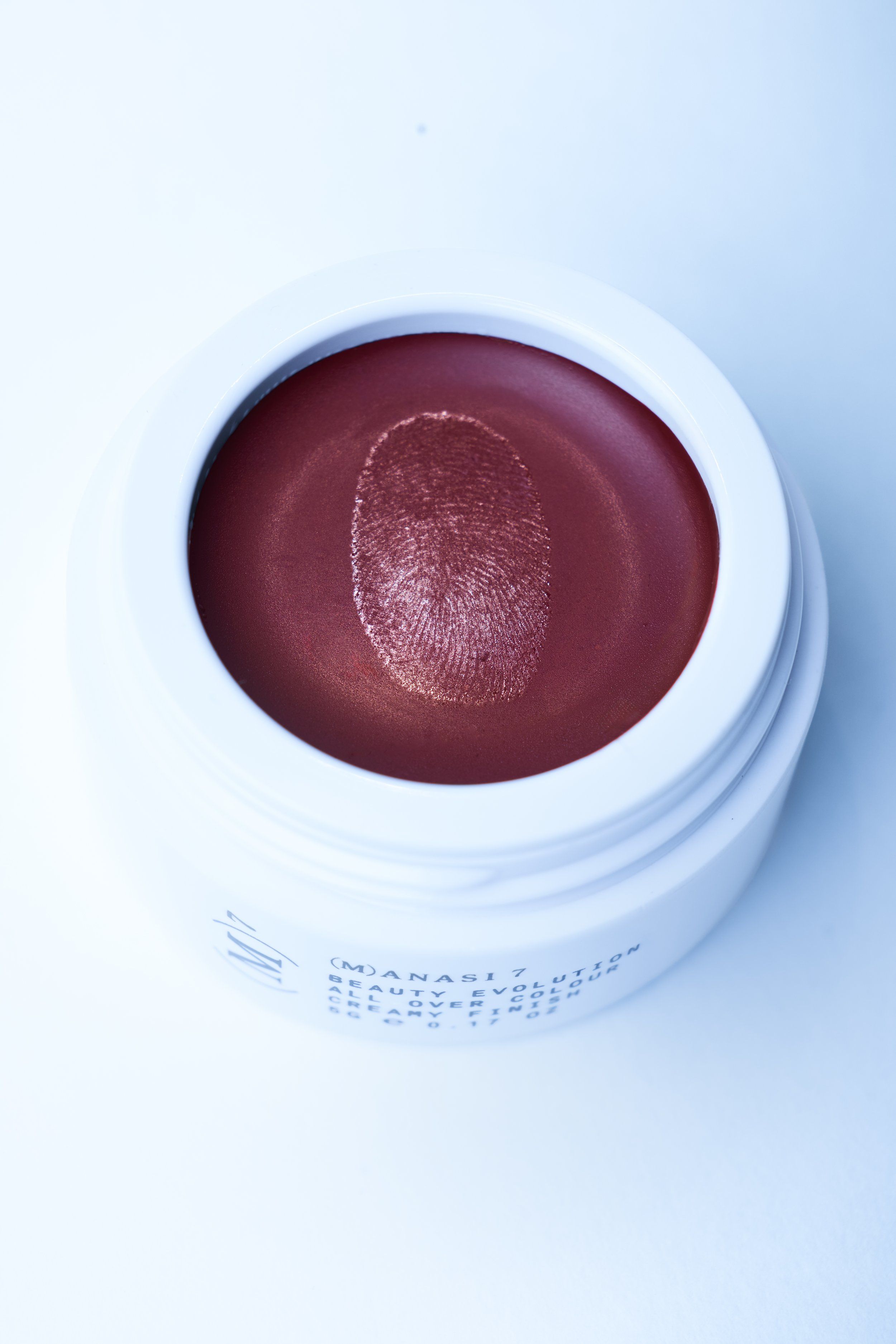 Beauty packshot of a white cream jar with a fingerprint imprint in the deep red product, on a pale blue background