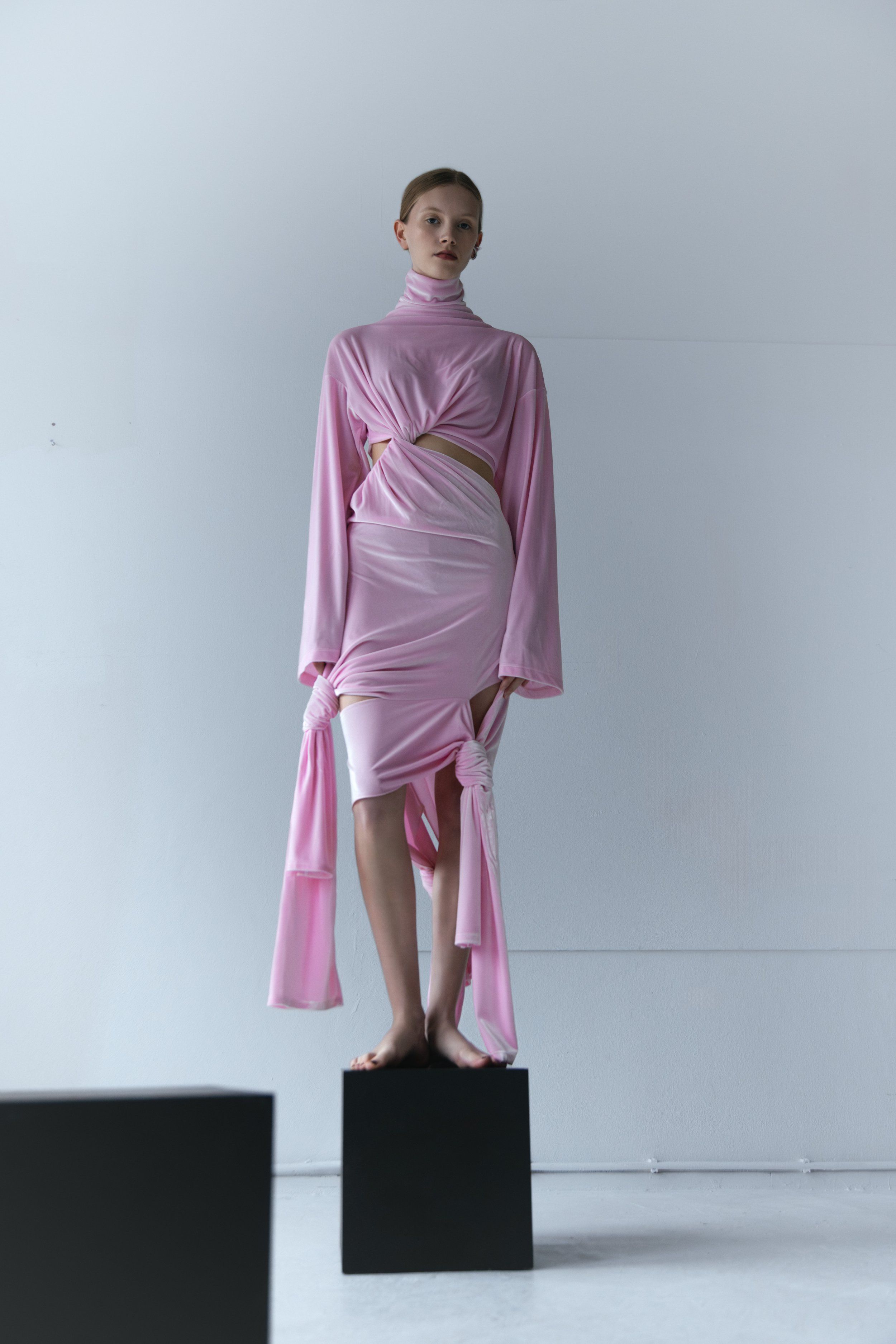 Model standing on a black cube in a pink velvet turtleneck top and skirt with long trailing ties, grey studio