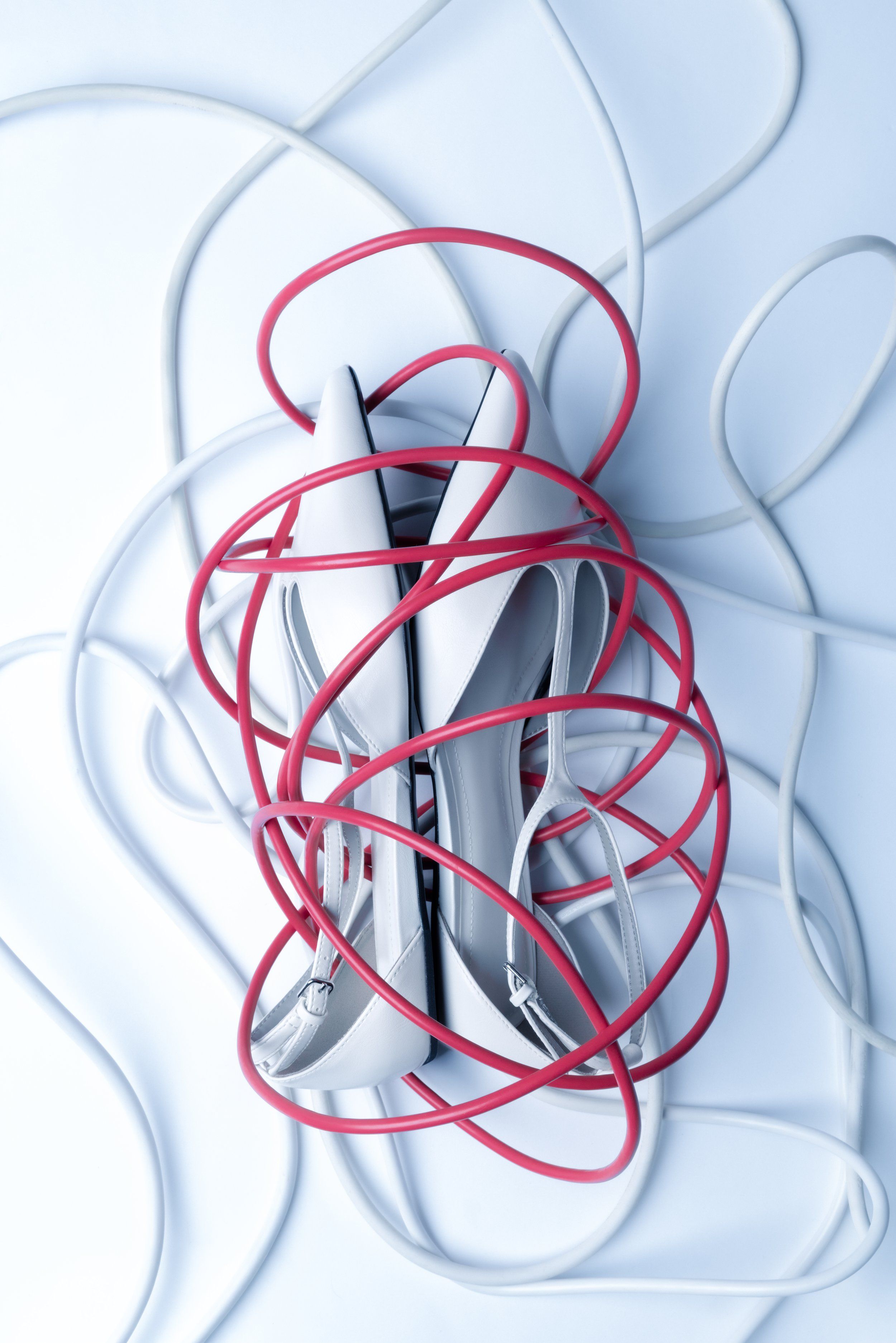 Product still life of pale pointed heels tangled in red and white cables on a light blue surface