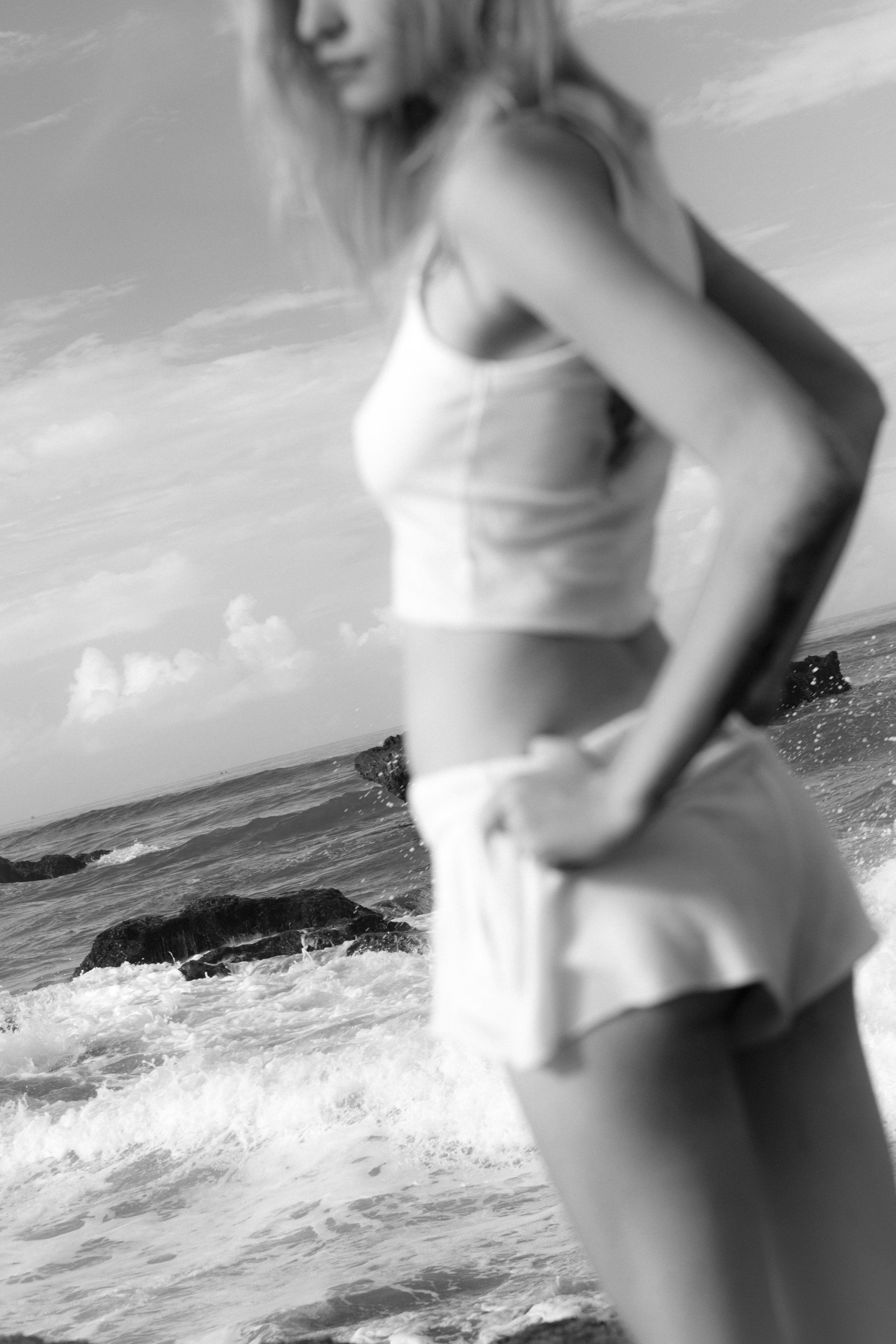 Black-and-white beach shot of a blurred model in a light crop top and shorts, waves and rocks behind