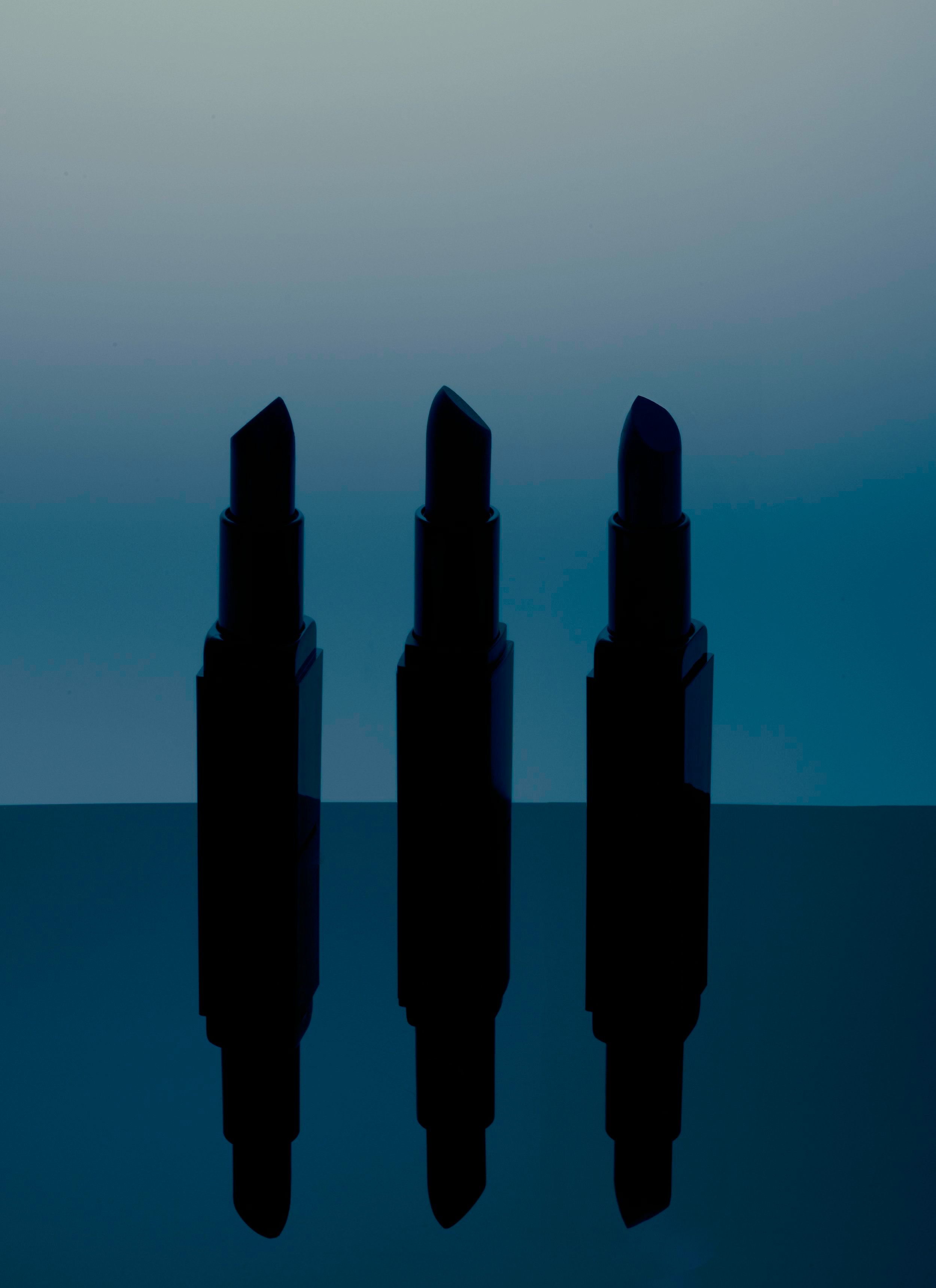 Three black lipsticks standing in silhouette against a dark blue gradient with mirrored reflection
