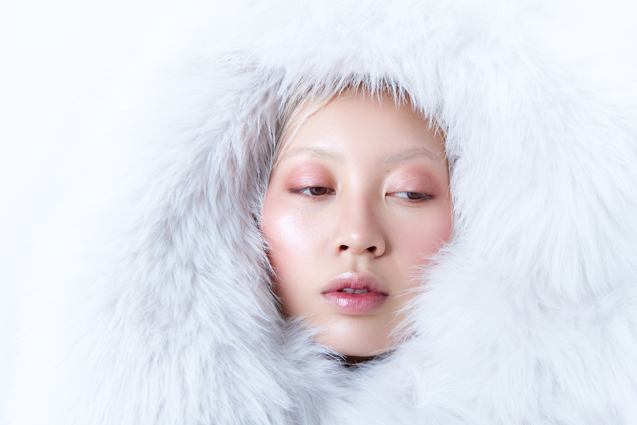 Beauty portrait of a model with pink-toned makeup framed by a white fluffy fur hood on a white background