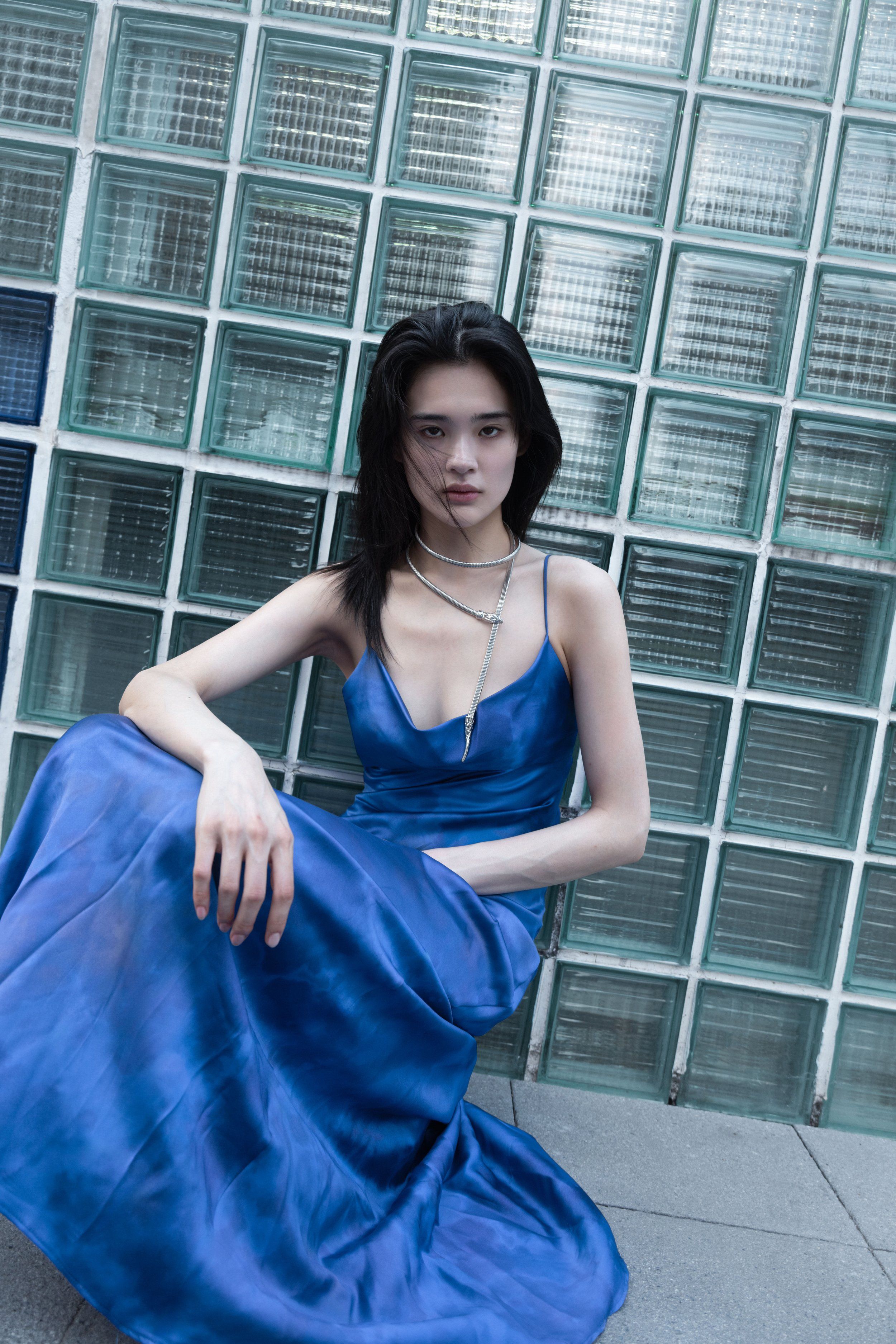 Model seated against a glass-block wall in a cobalt blue satin slip dress with layered necklaces