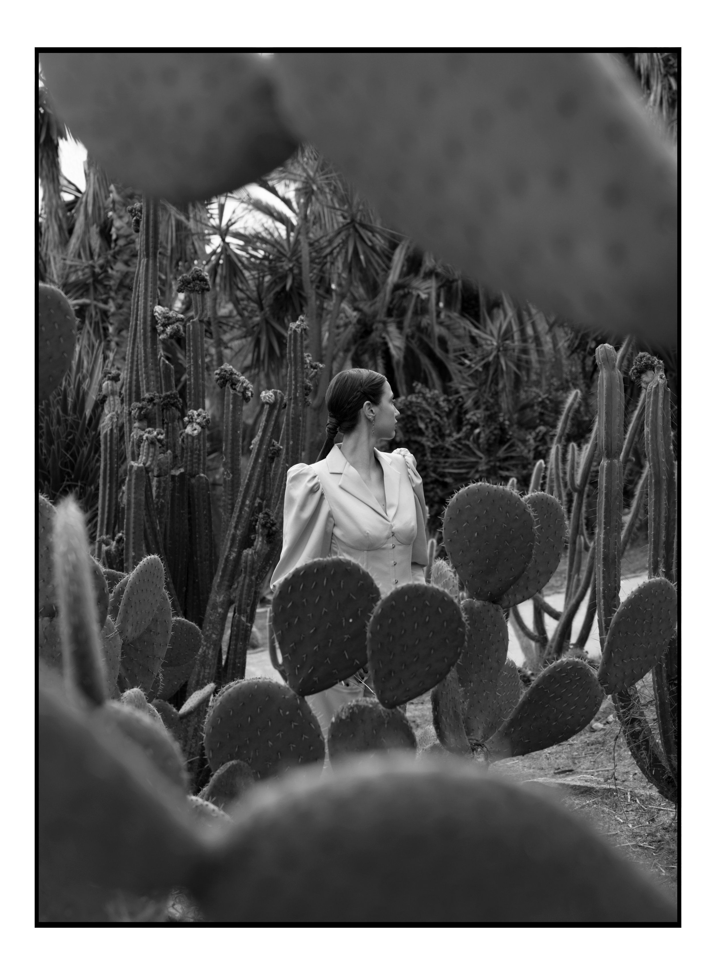 Black-and-white image of a woman in a light blazer seen through cactus leaves in a botanical garden, black border