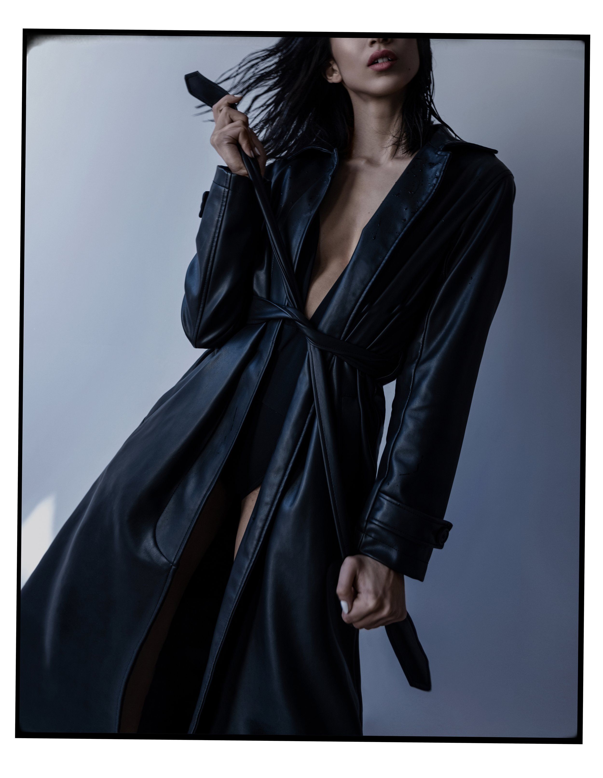 Model in a long black leather trench coat with belt in hand, grey studio wall, framed film-style border