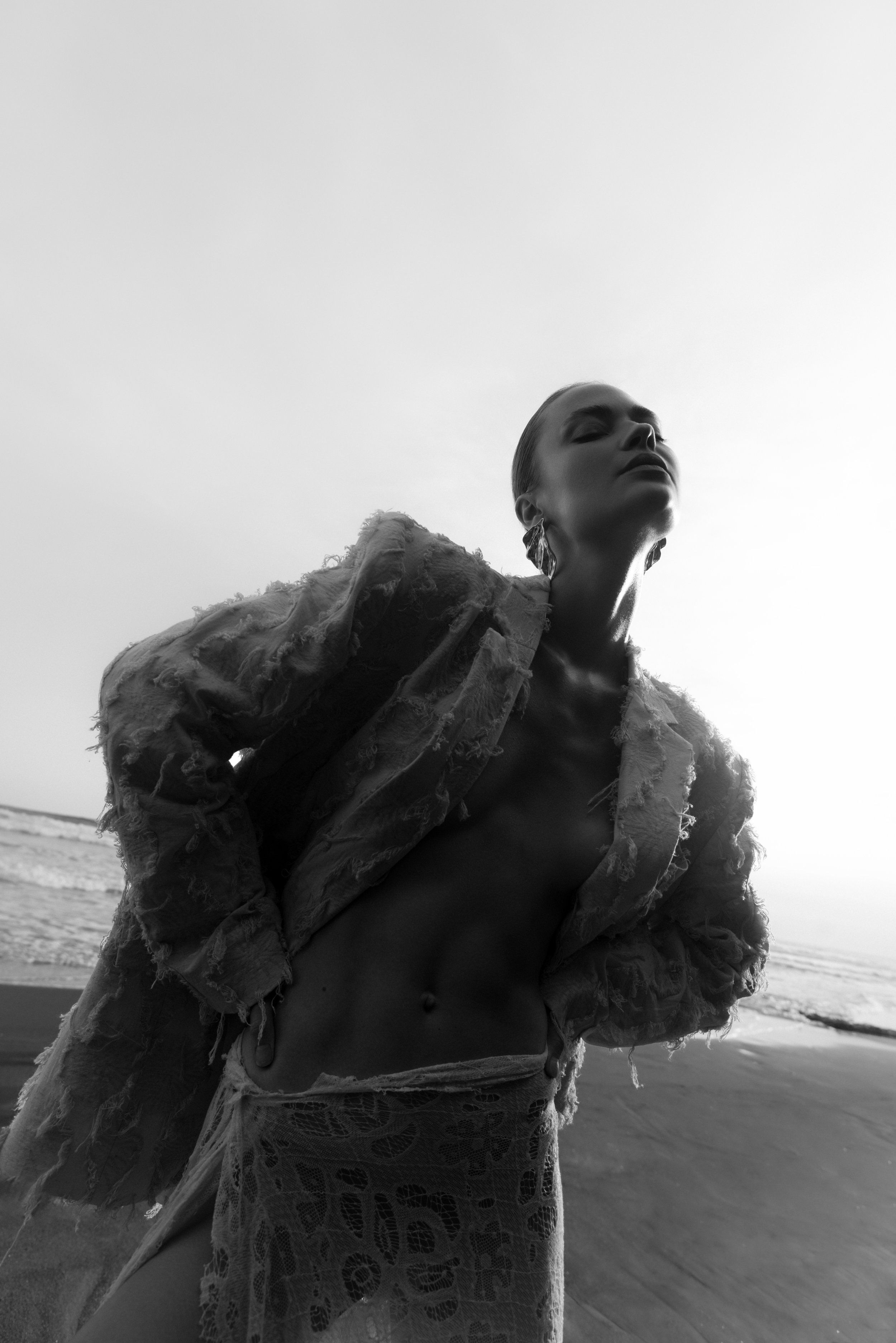 Black-and-white beach portrait of a model with head tilted back in a frayed textured jacket and lace skirt