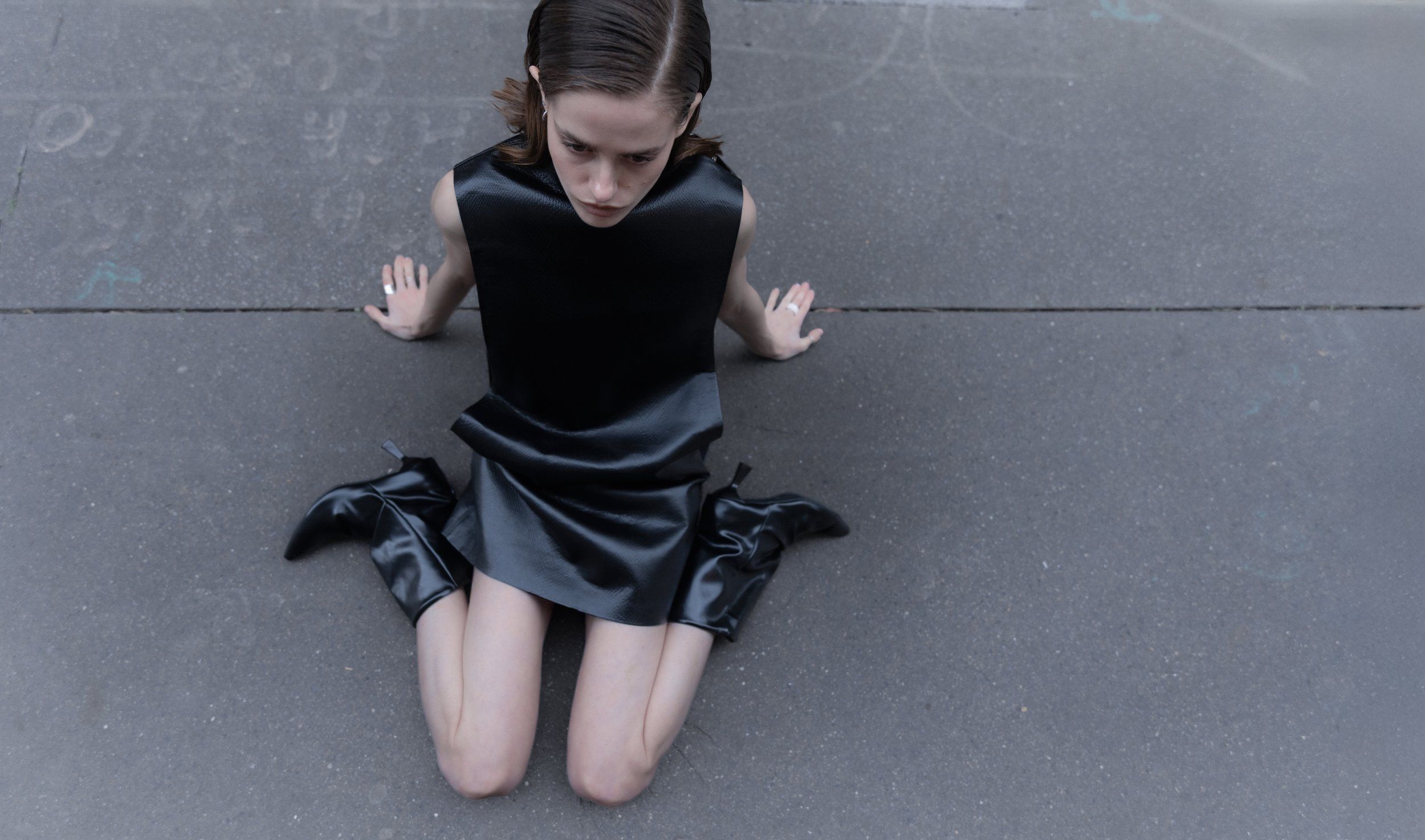 Model kneeling on grey asphalt in a black leather mini dress and black ankle boots, shot from above