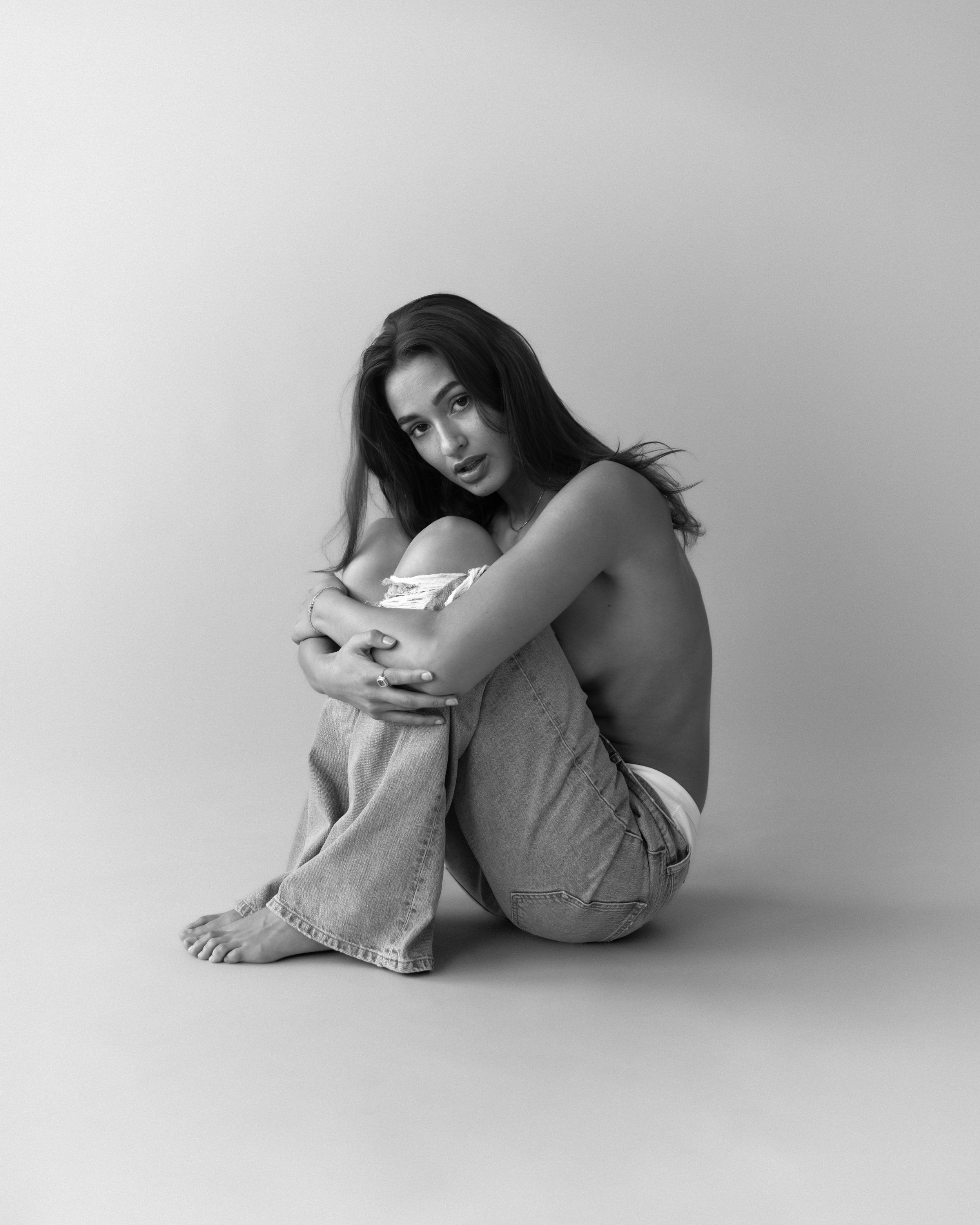 Black-and-white studio portrait of a model seated on the floor hugging her knees in wide-leg jeans, grey backdrop