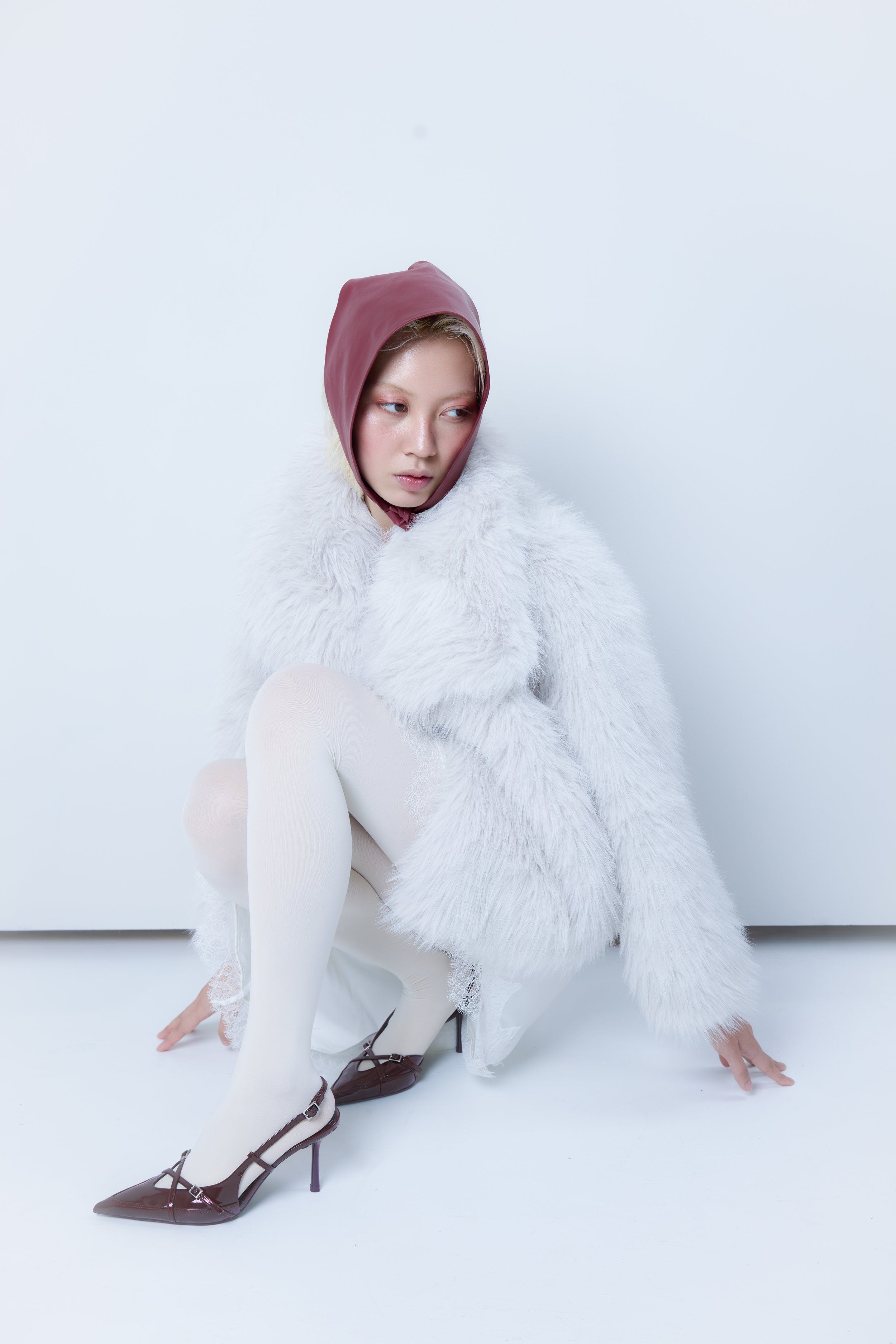 Model crouching on a white floor in a burgundy hood, white faux-fur coat, white tights and maroon slingback heels