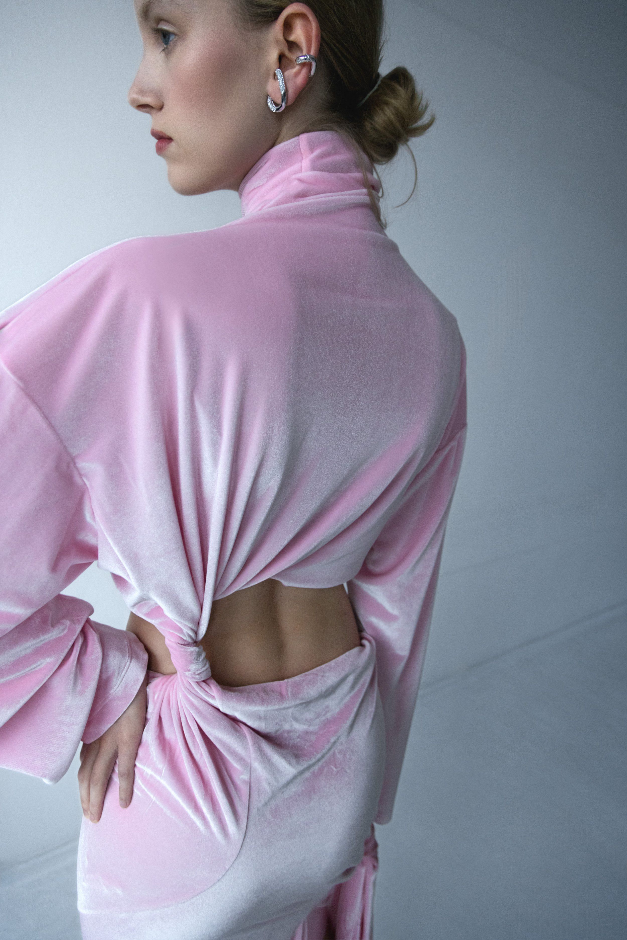 Rear three-quarter view of a model in a pink velvet turtleneck with cut-out back, silver ear cuffs, grey backdrop