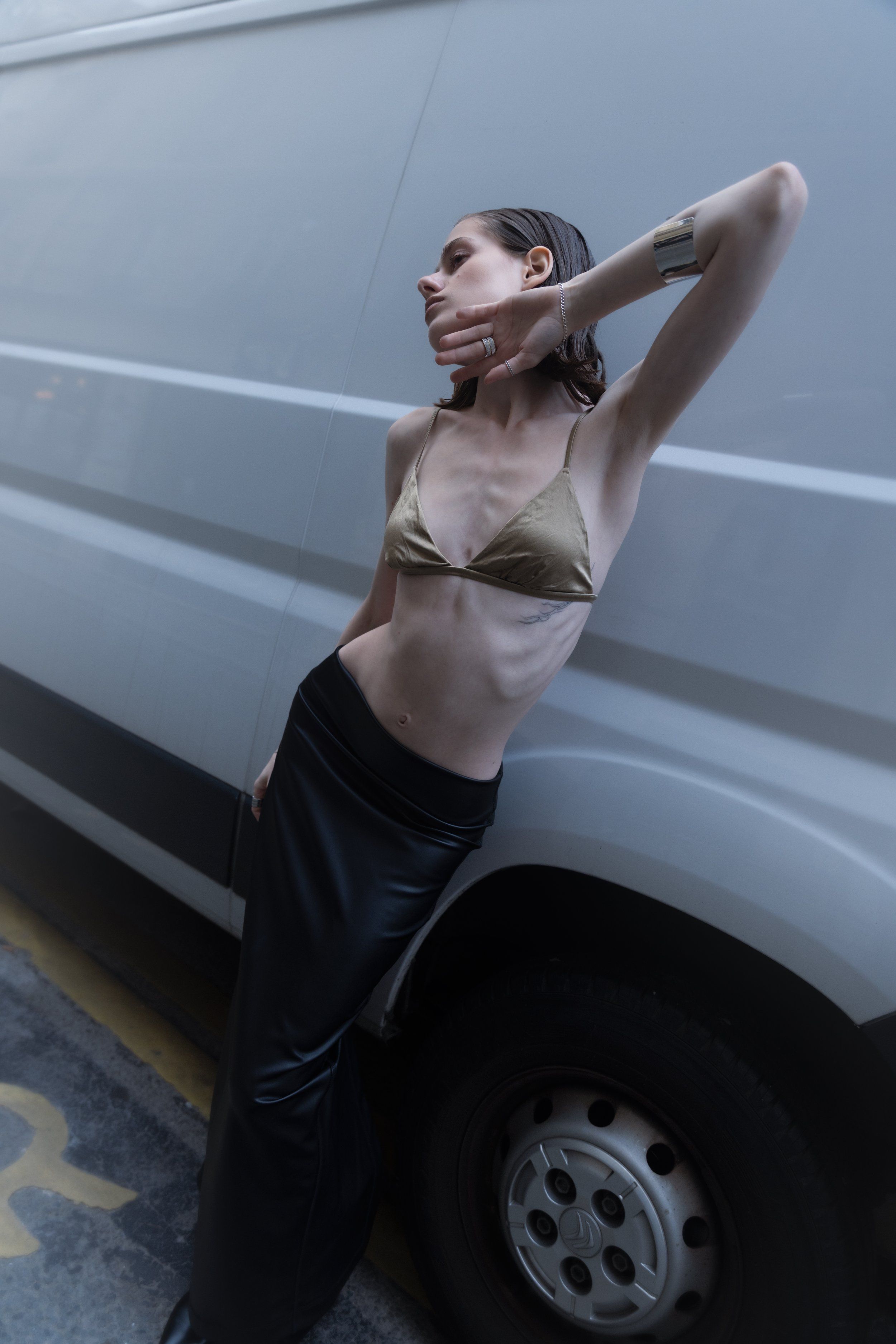 Model leaning against a grey van in a satin bralette and black leather skirt, one arm raised, street setting