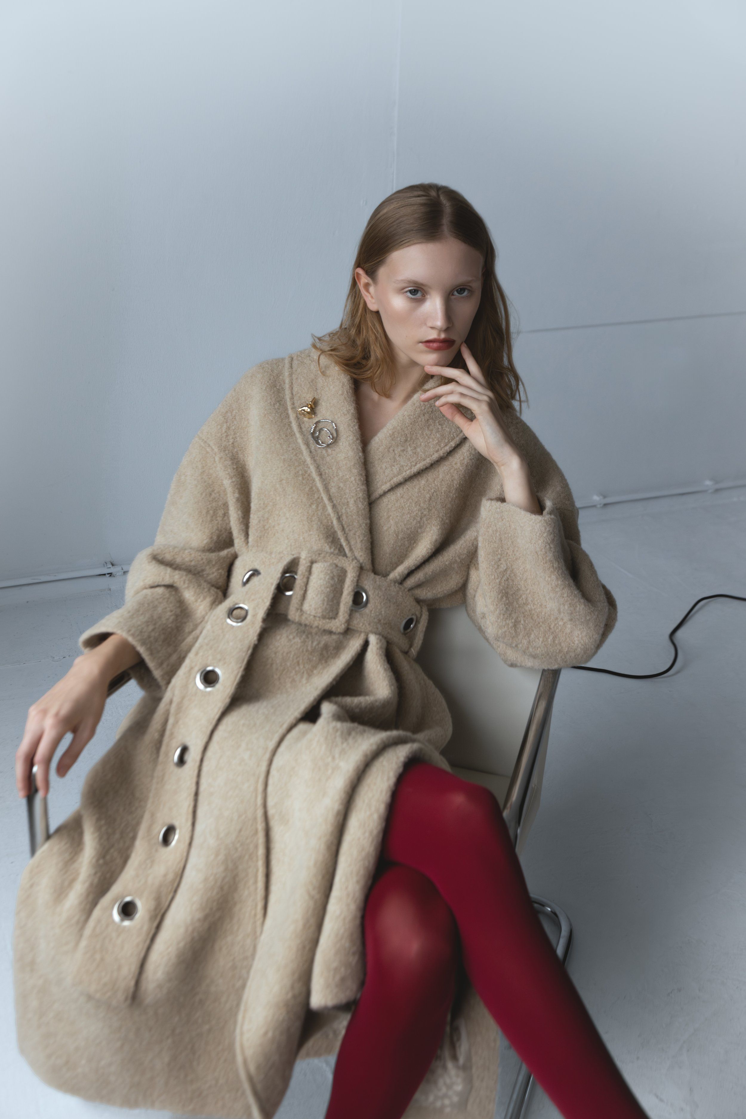 Model seated on a chair in a belted beige wool coat with eyelets and red tights, grey studio backdrop