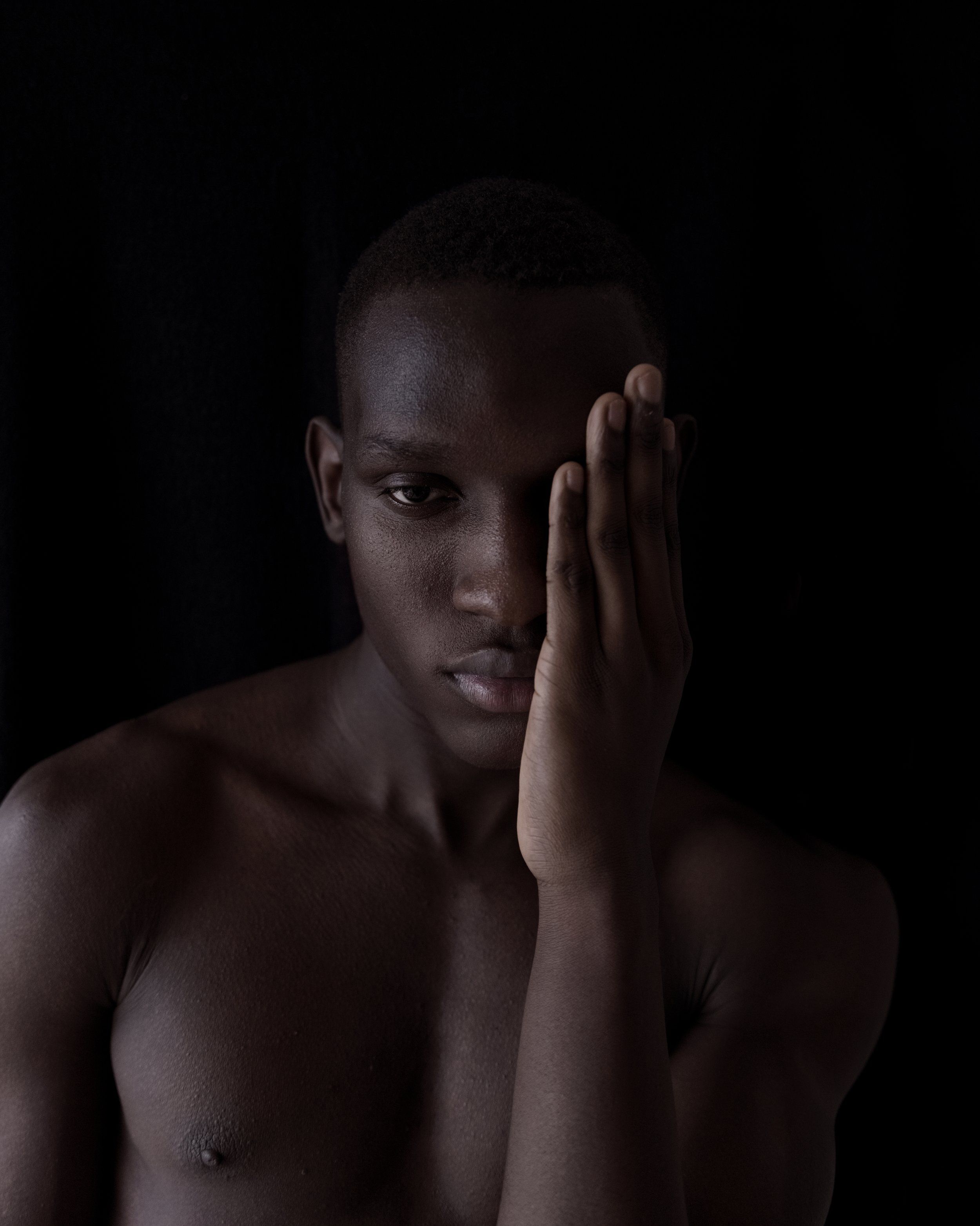 Moody low-key portrait of a young man with a hand covering half his face, dark background, bare shoulders