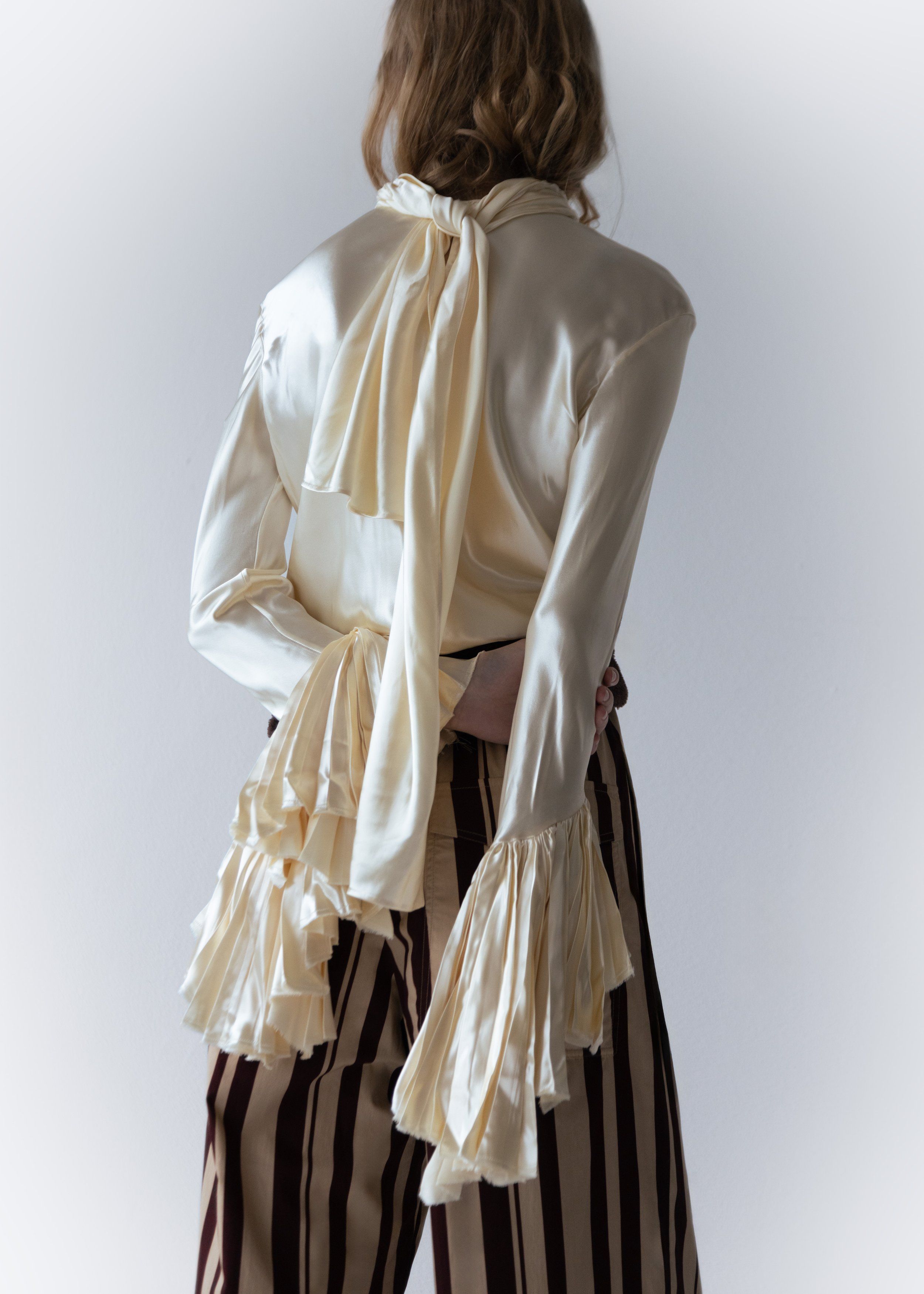 Model seen from behind in a cream satin ruffled blouse with long tie and striped trousers, soft grey background