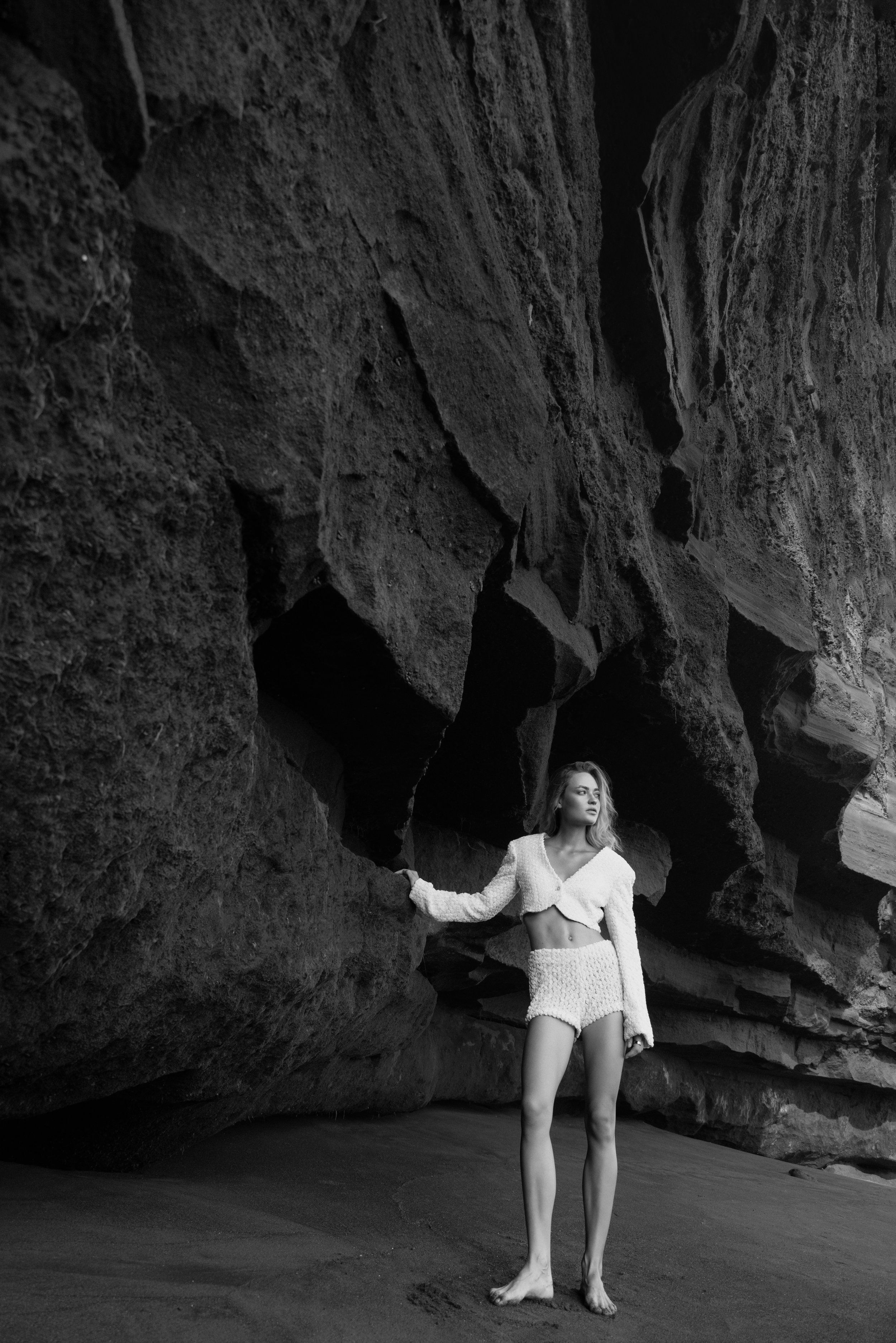 Black-and-white image of a model in white shorts and cropped jacket standing before a dark cave wall on the beach