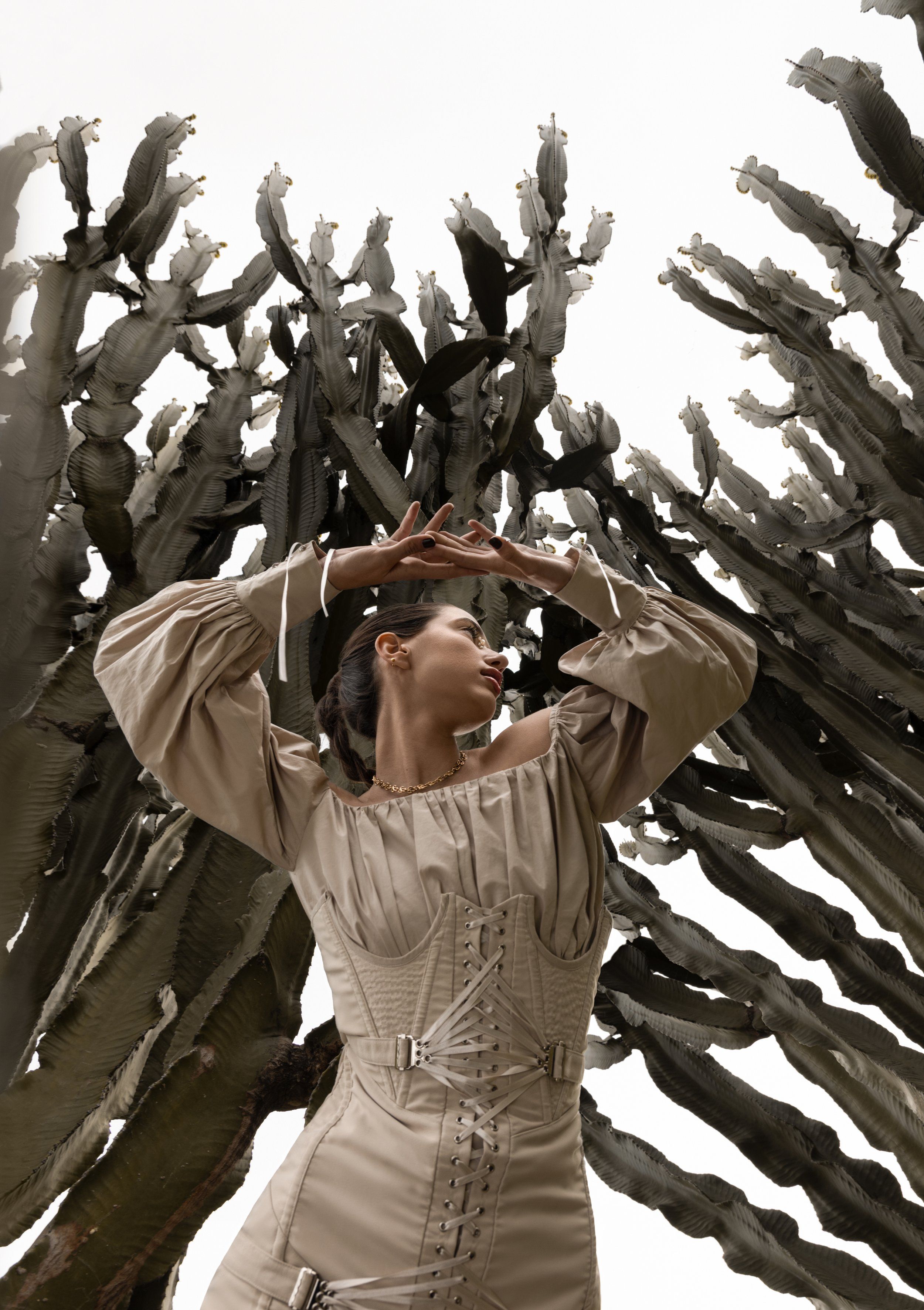 Model in a beige corseted top with puffed sleeves raising her arms among tall cactus branches against a pale sky