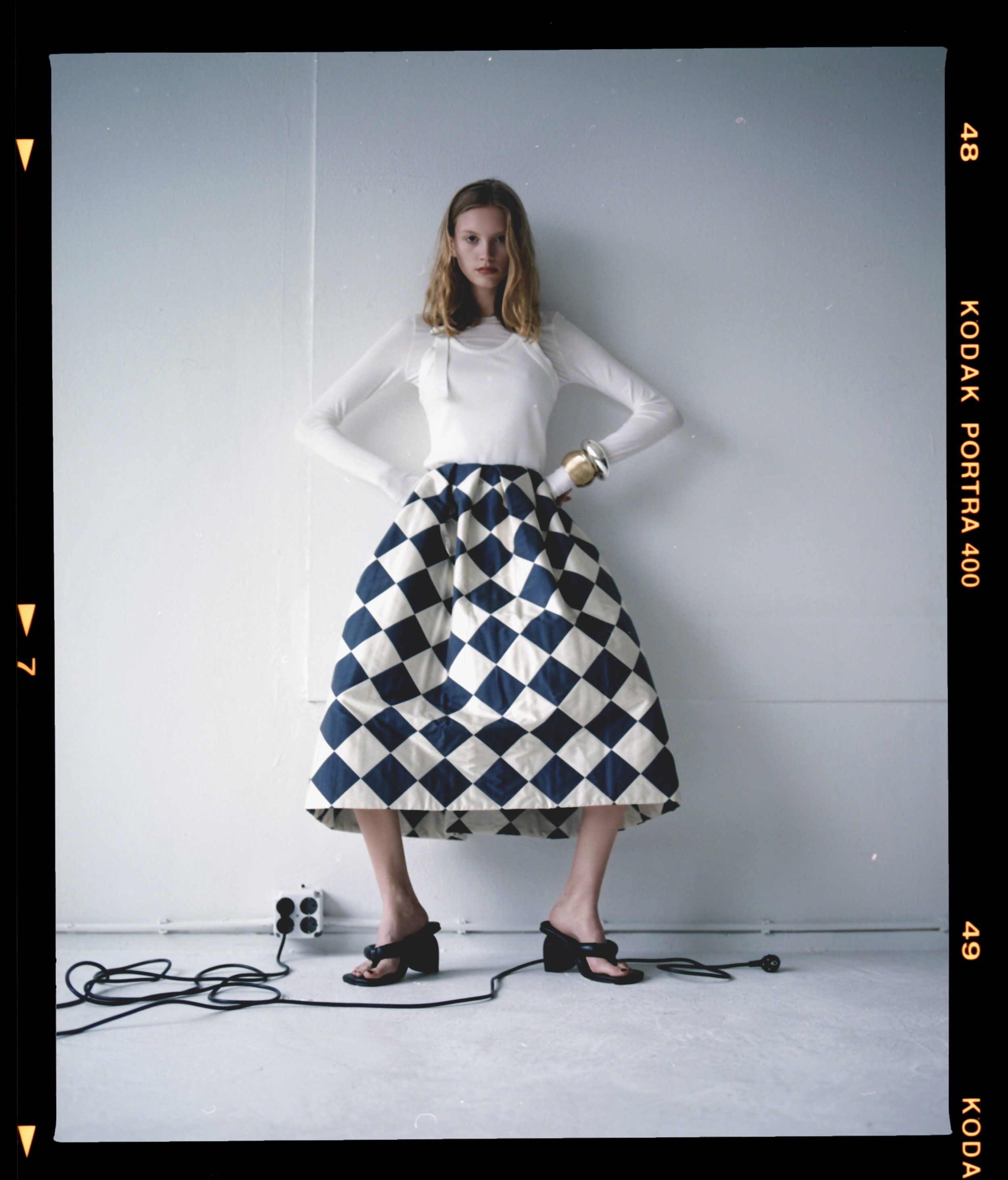 Film-frame portrait of a model in a navy and cream diamond-pattern skirt, white top and black sandals, Kodak Portra border