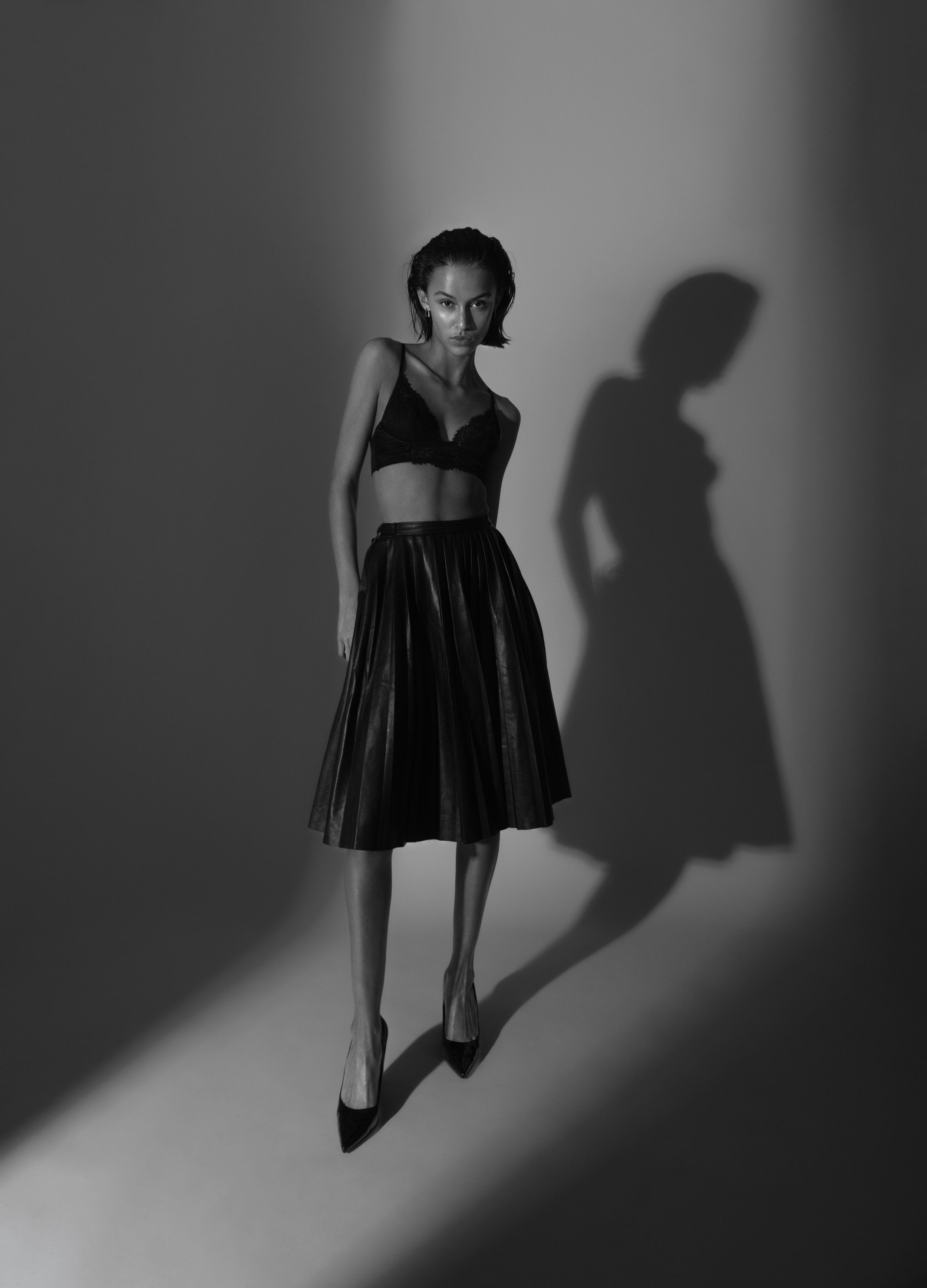 Black-and-white studio portrait of a model in black bra top and leather skirt, strong spotlight casting a long shadow