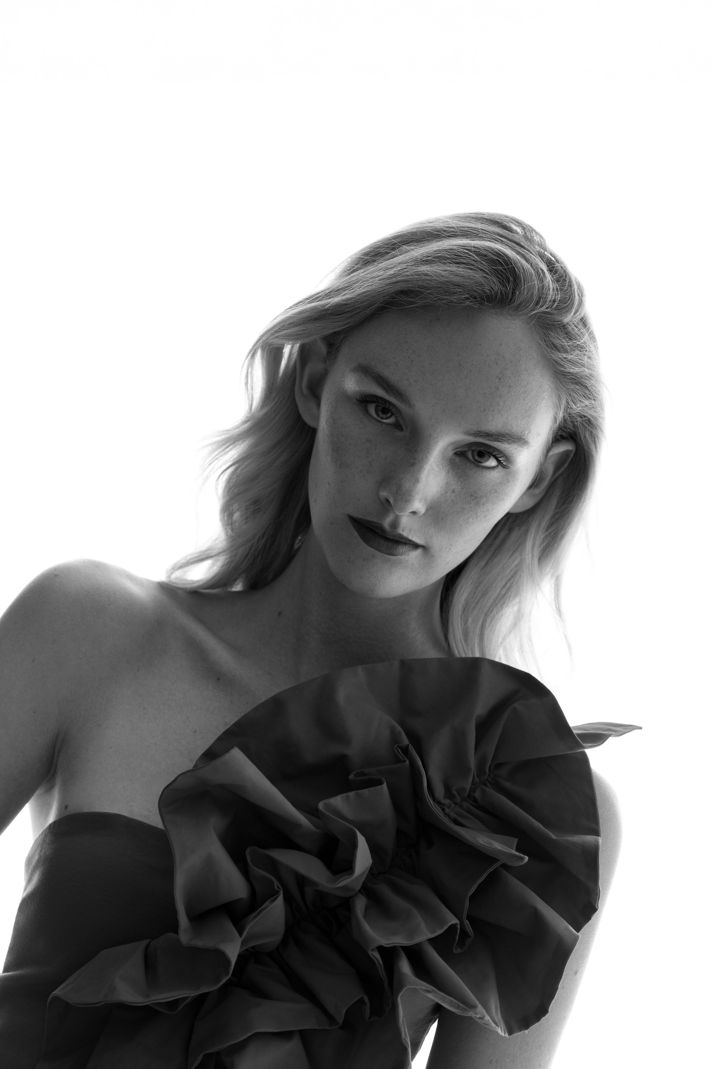 Black-and-white portrait of a blonde model in a strapless dress with a large ruffled neckline, bright white background