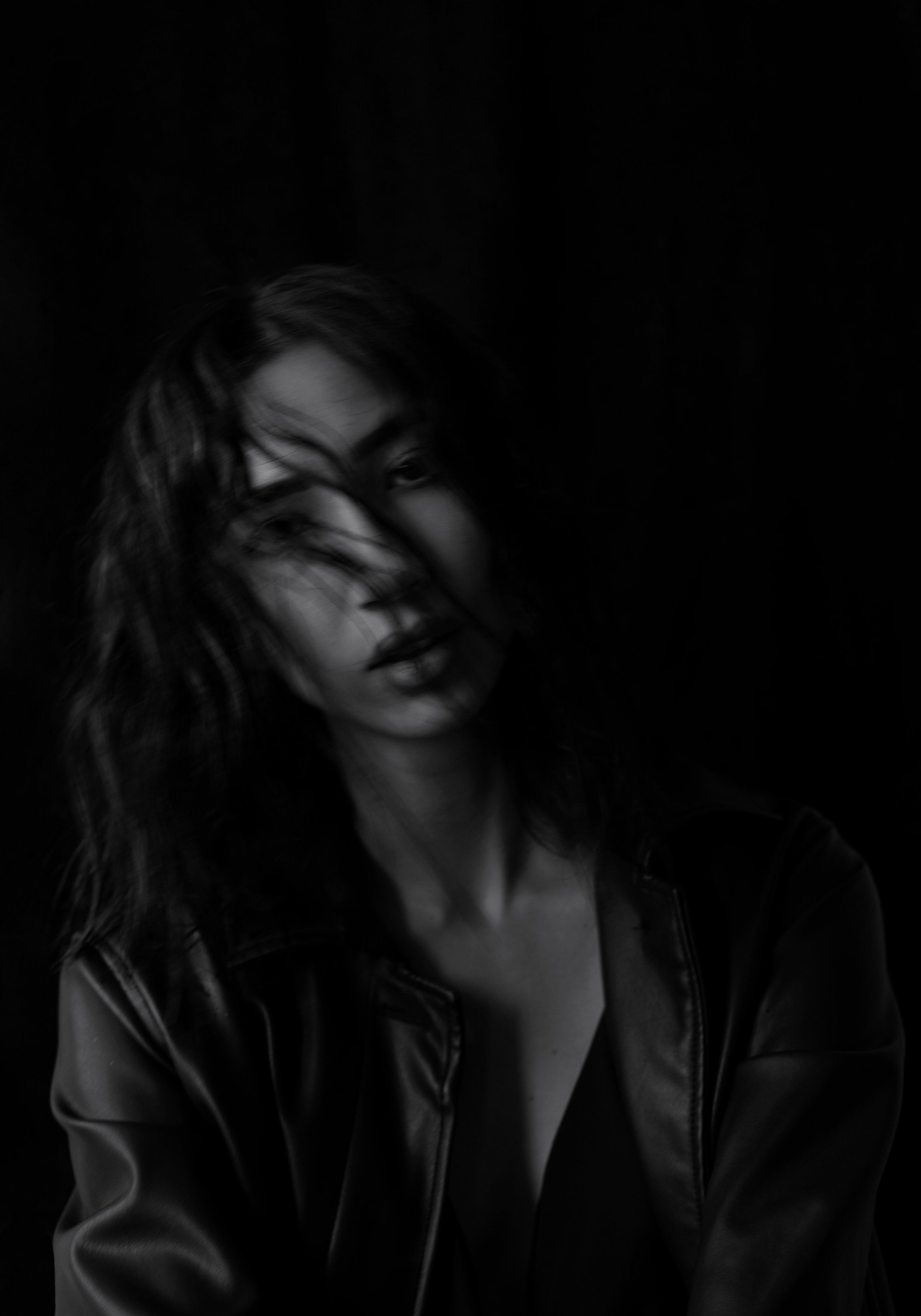 Low-key black-and-white portrait of a model with hair across her face in a leather jacket, deep shadows