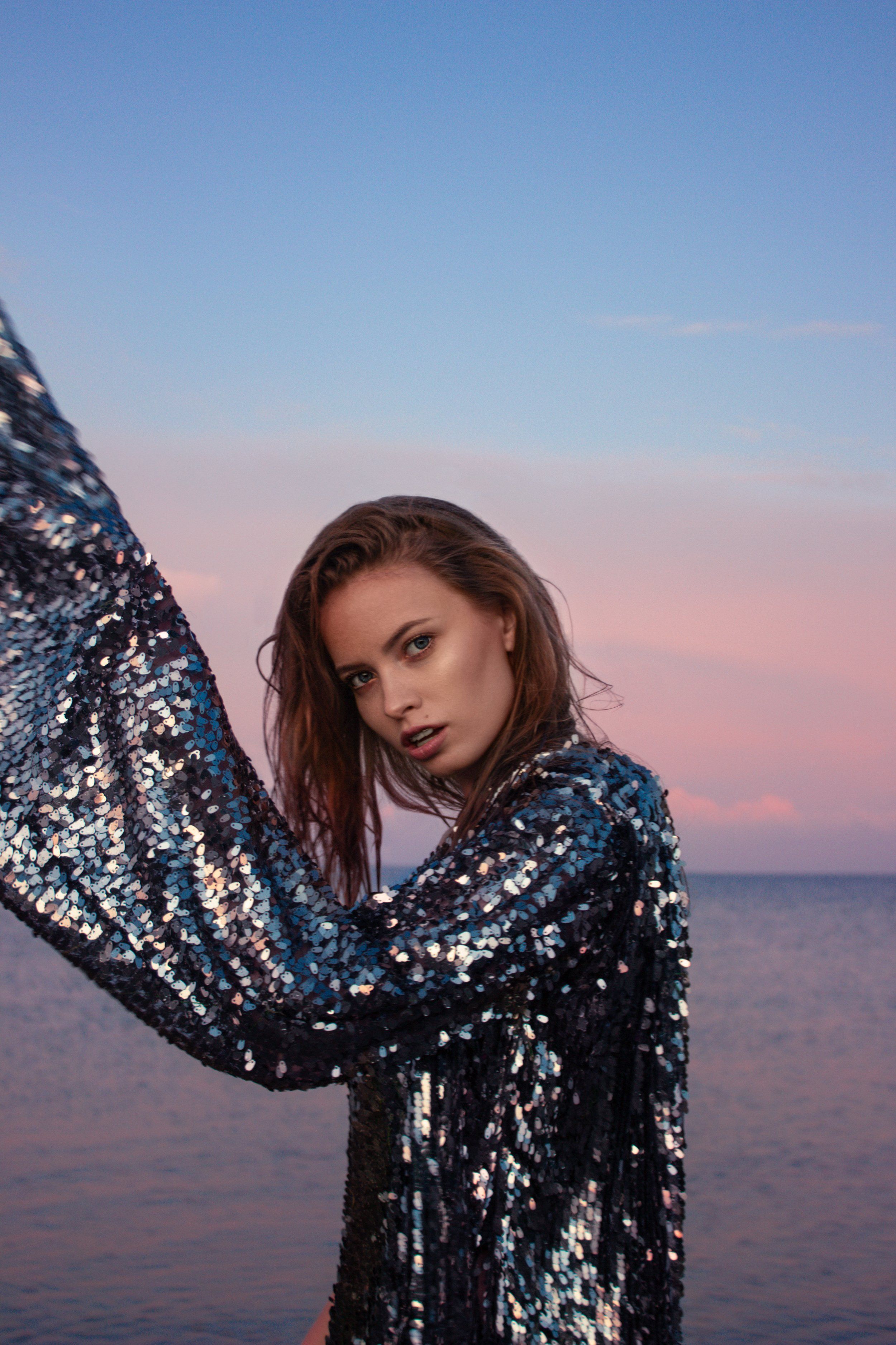 Model in a sequined jacket against a pink and blue dusk sky over the sea, one arm stretched out
