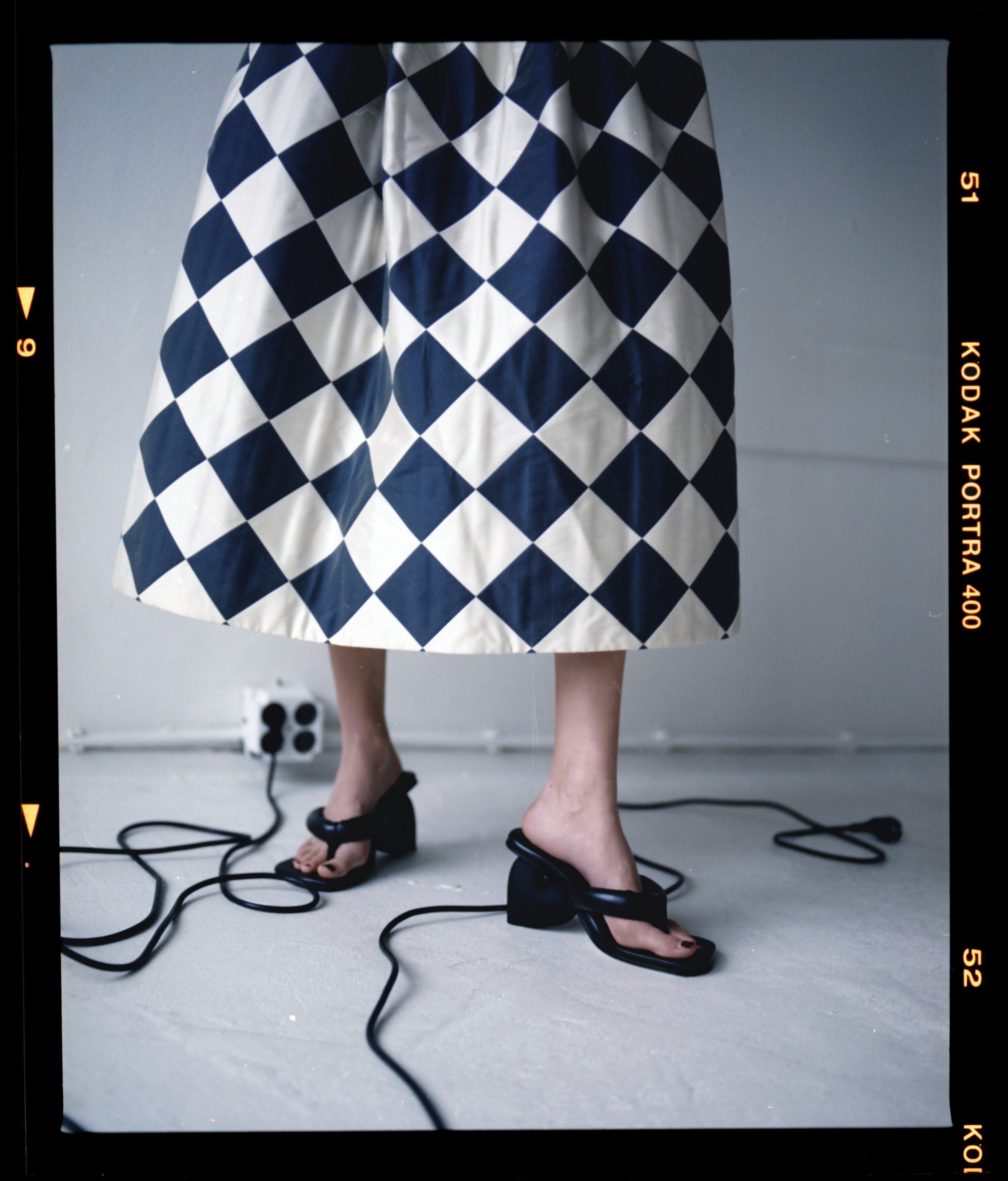 Film-frame detail of a diamond-pattern skirt hem and black platform sandals with cables on the floor, Kodak Portra border