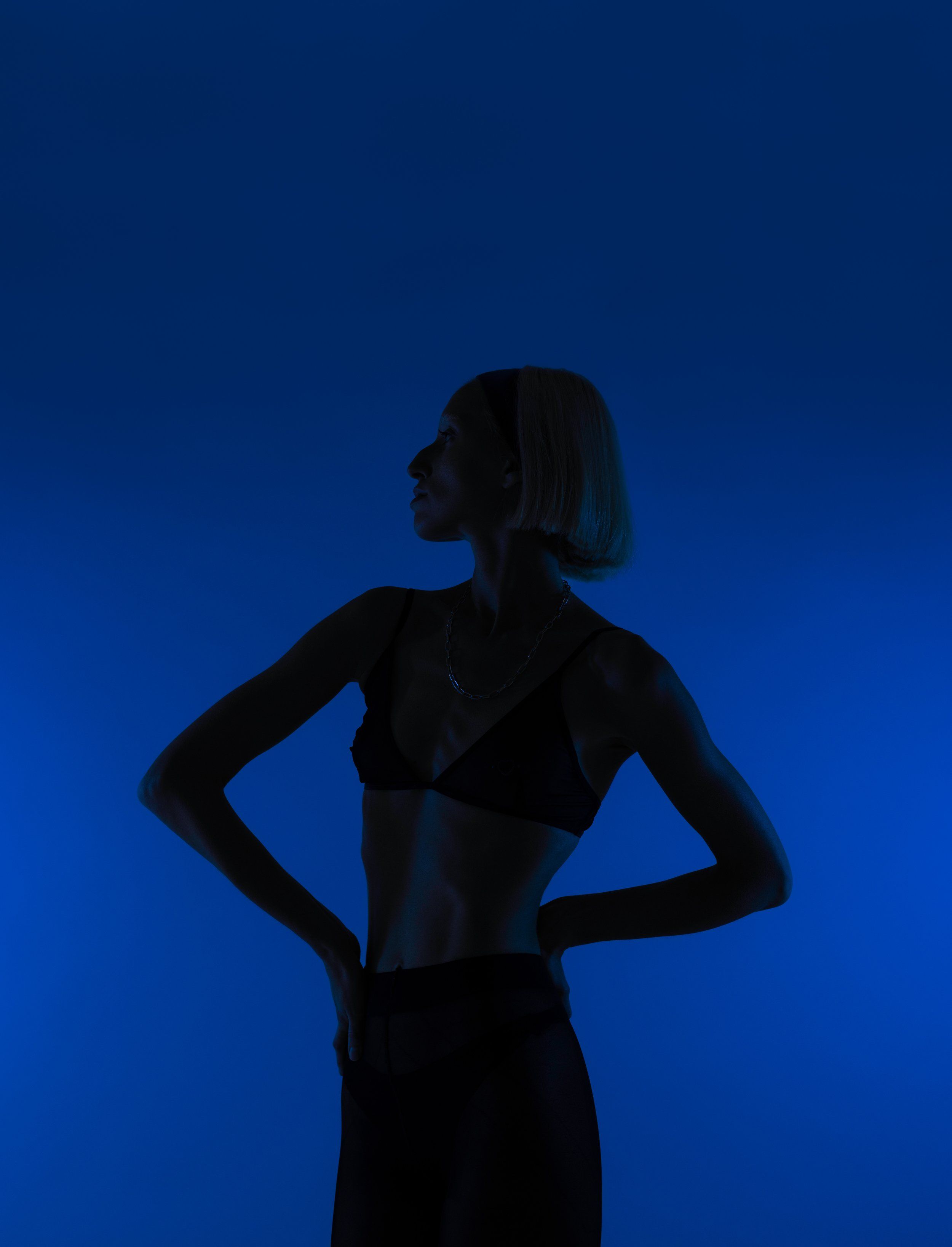 Backlit silhouette of a model in a black bra top with hands on hips against a saturated blue background