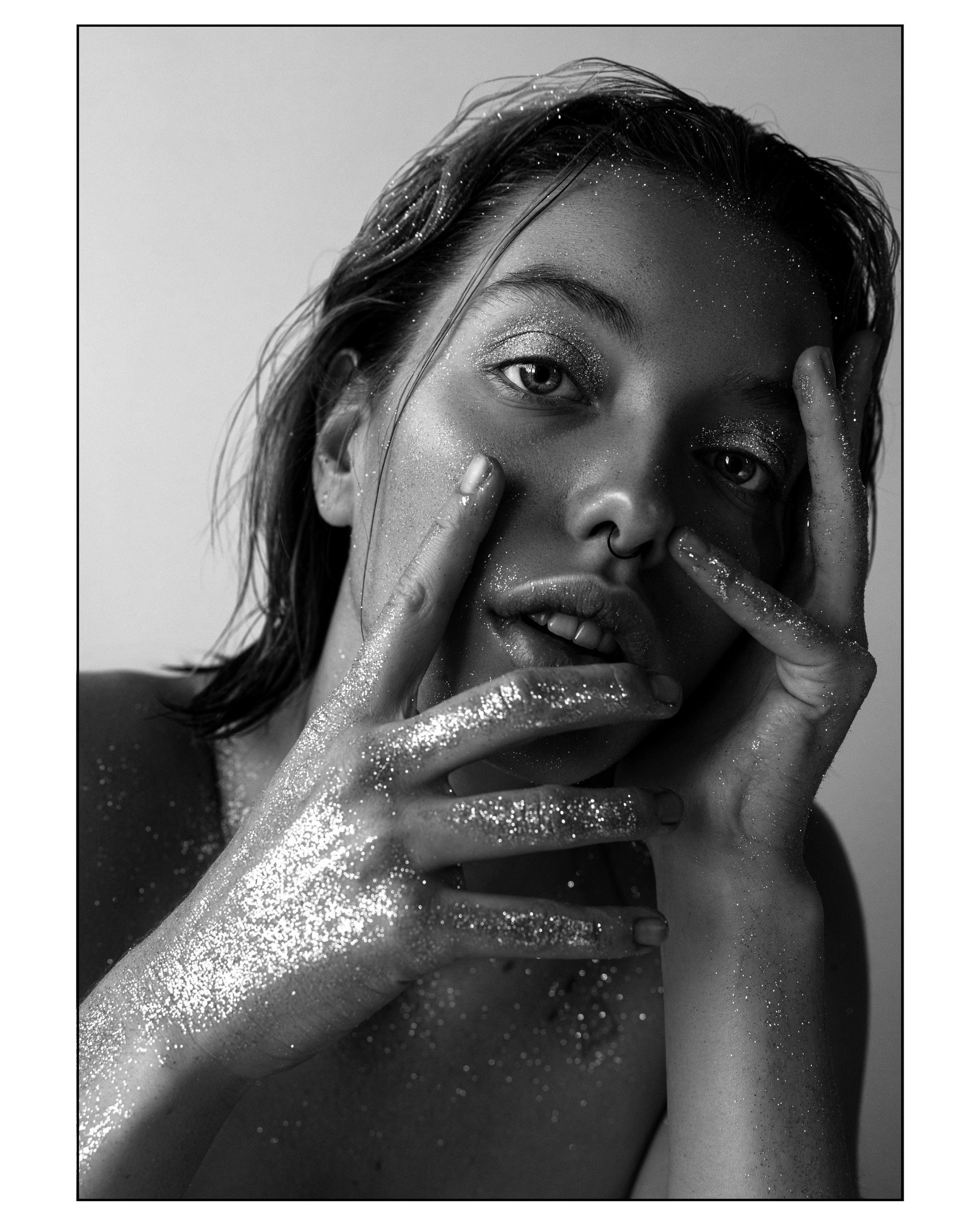 Black-and-white portrait of a model with glitter on her hands and face, fingers framing her face, black border